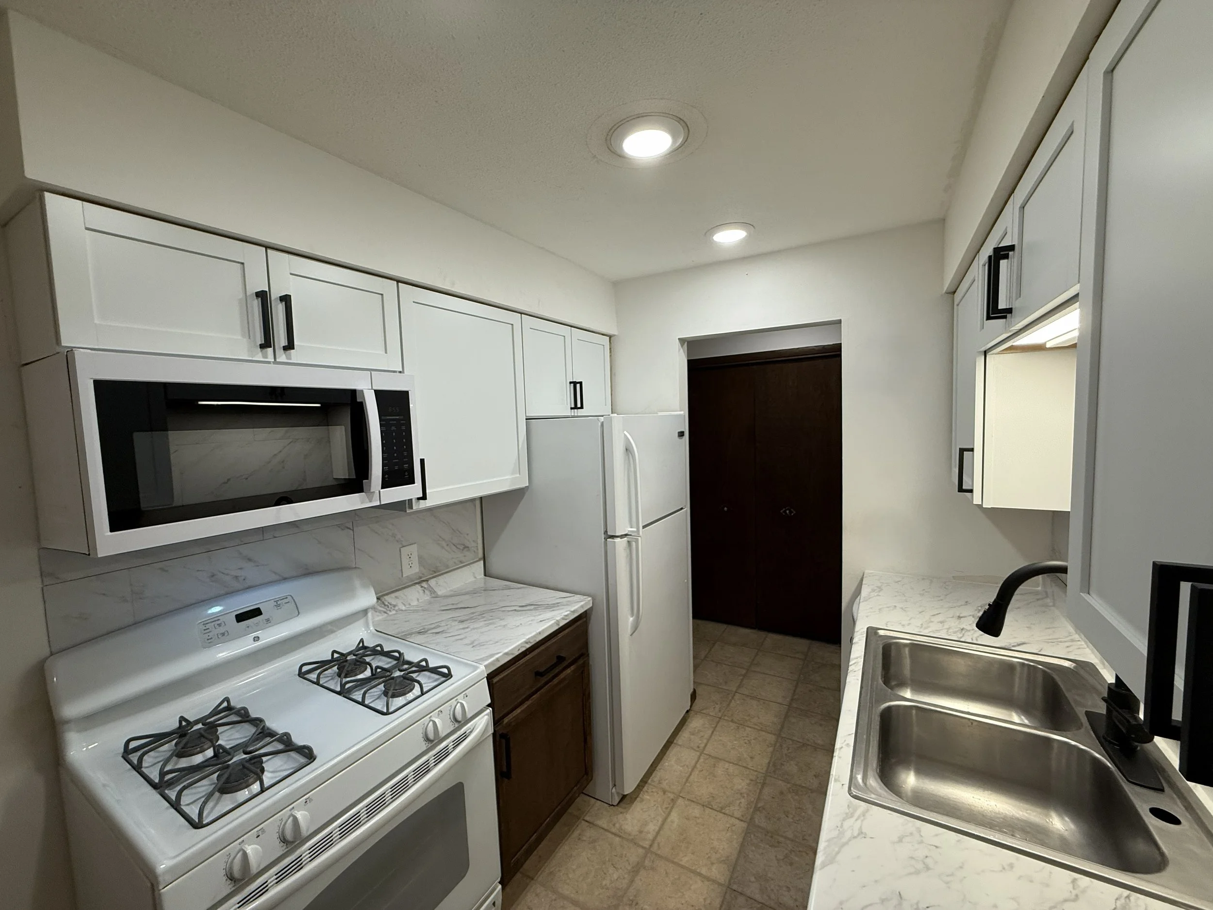 A kitchen with white cabinets, a stove, microwave, refrigerator, and a double sink, with tiled flooring and ceiling lights.
