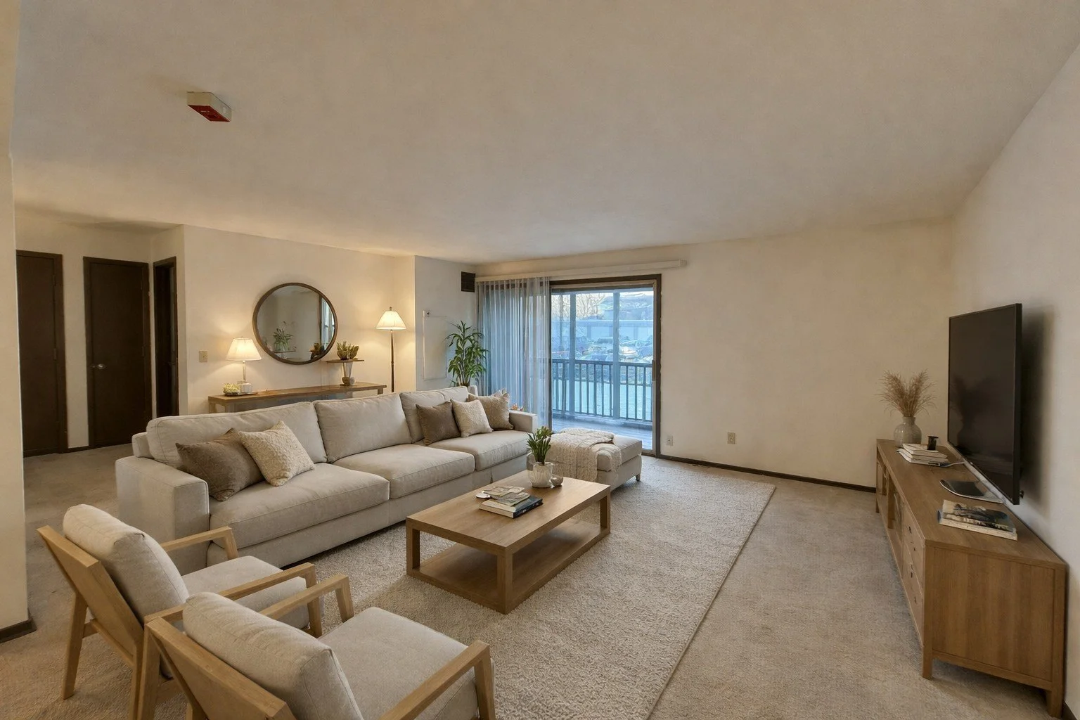 Living room with beige sofa, two cream armchairs, wooden coffee table, TV on wooden stand, sliding glass door leading to balcony, and decorative lamps and plants.