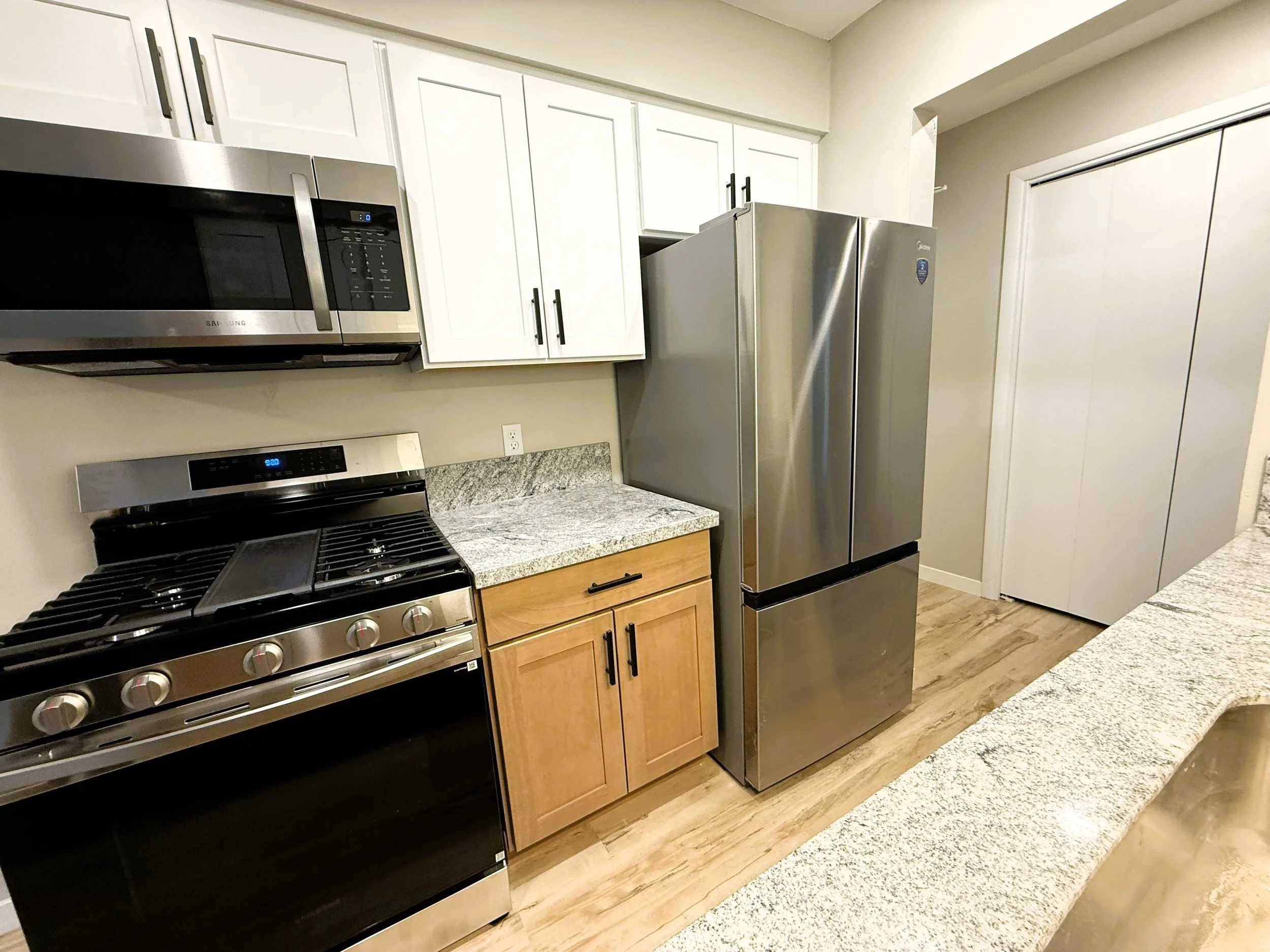 Kitchen with stainless steel microwave, refrigerator, oven, and granite countertops. White cabinets with black handles are above the appliances. Light-colored flooring.