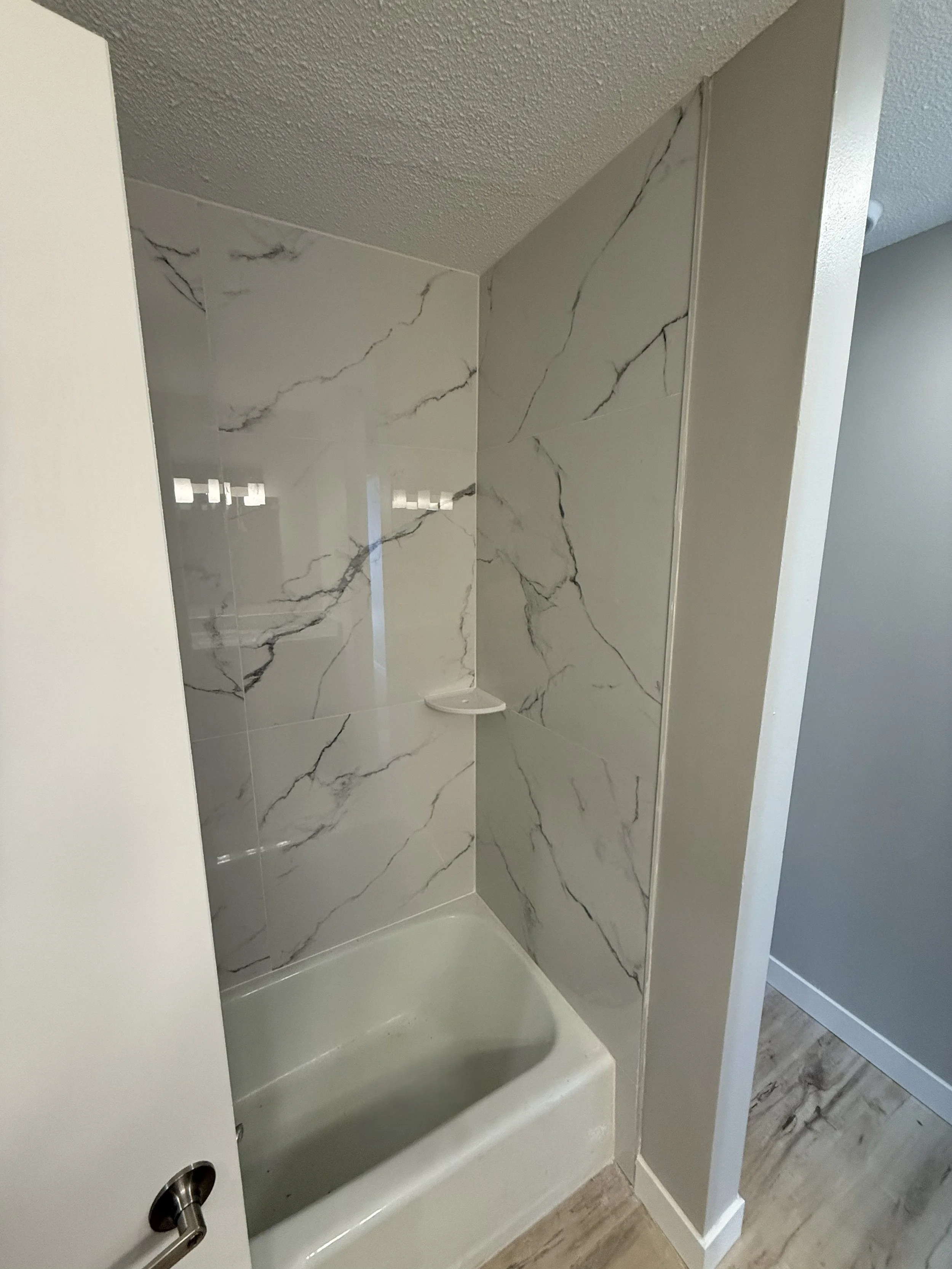 A shower with marble tiled walls and a shower tray. Part of the bathroom with a door handle visible in the foreground.