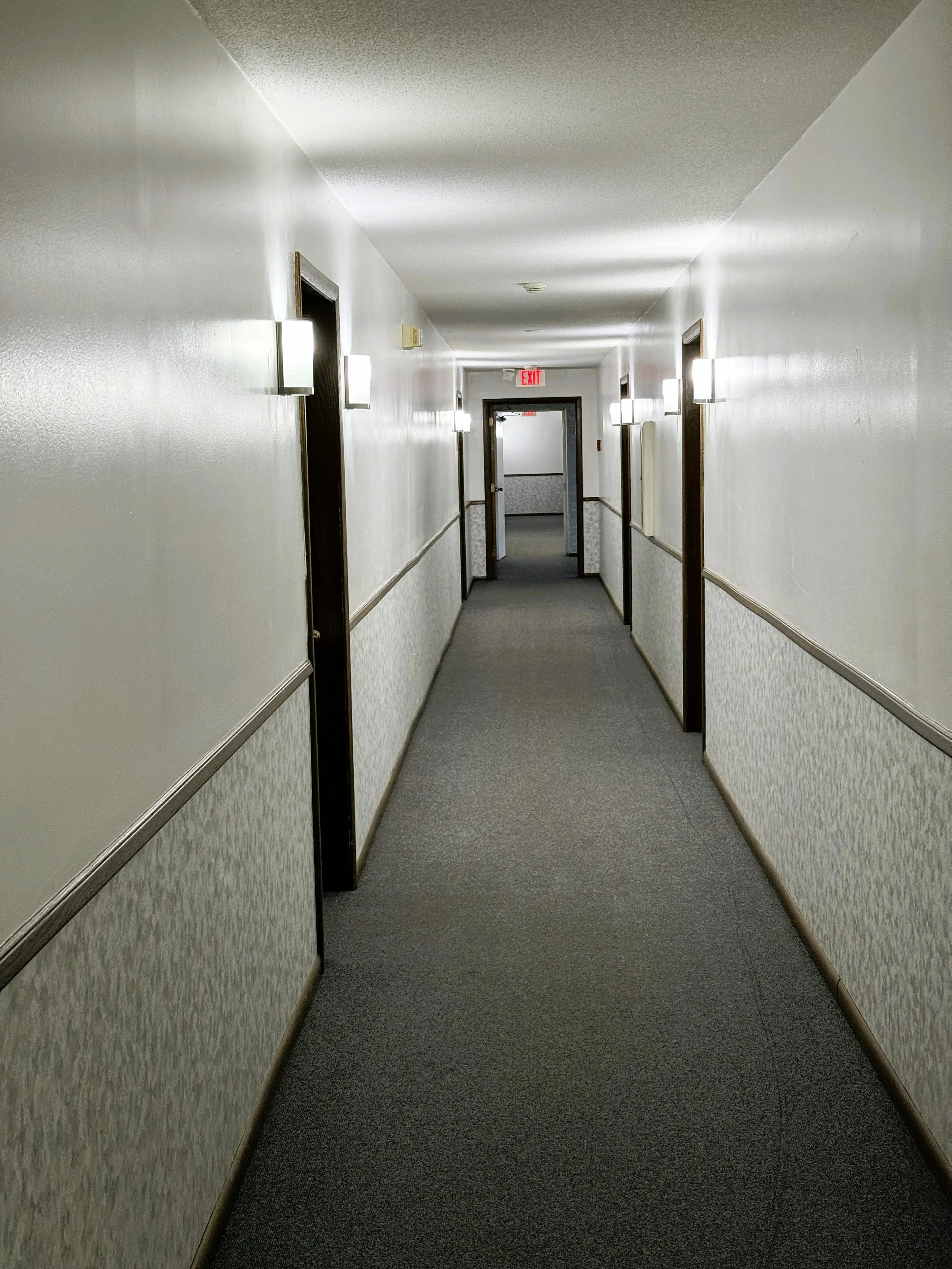 Empty hotel corridor with multiple doorways, wall-mounted lights, and an exit sign at the end.