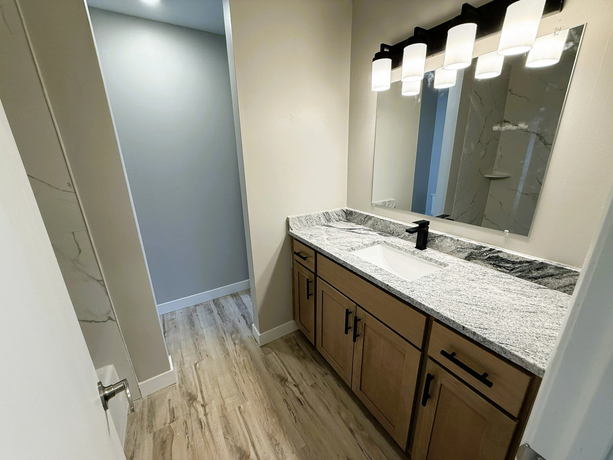 A bathroom vanity with a granite countertop and a rectangular mirror above it. Four cylindrical light fixtures are mounted above the mirror. The vanity has multiple wooden cabinets with black handles. The bathroom has light-colored walls and wood-loo