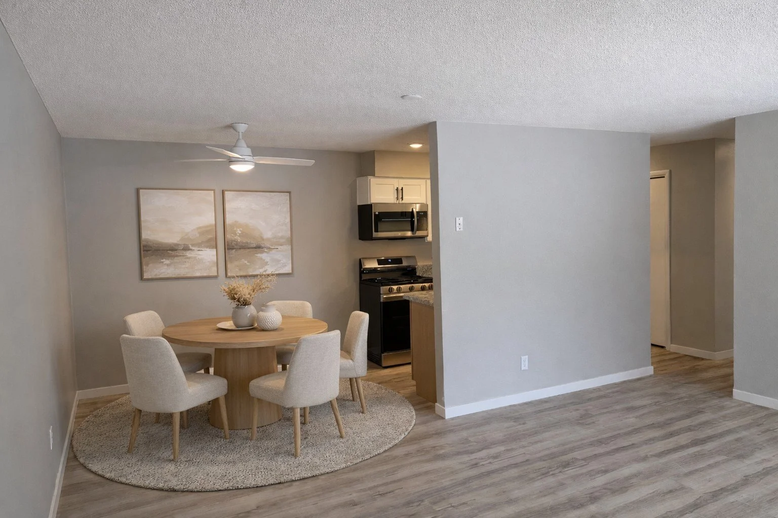 Dining area with a round wooden table, six beige upholstered chairs, and decorative vases on top. A textured area rug is underneath the table. In the background is a kitchen with a black stove and stainless steel microwave, white cabinets, and gray w