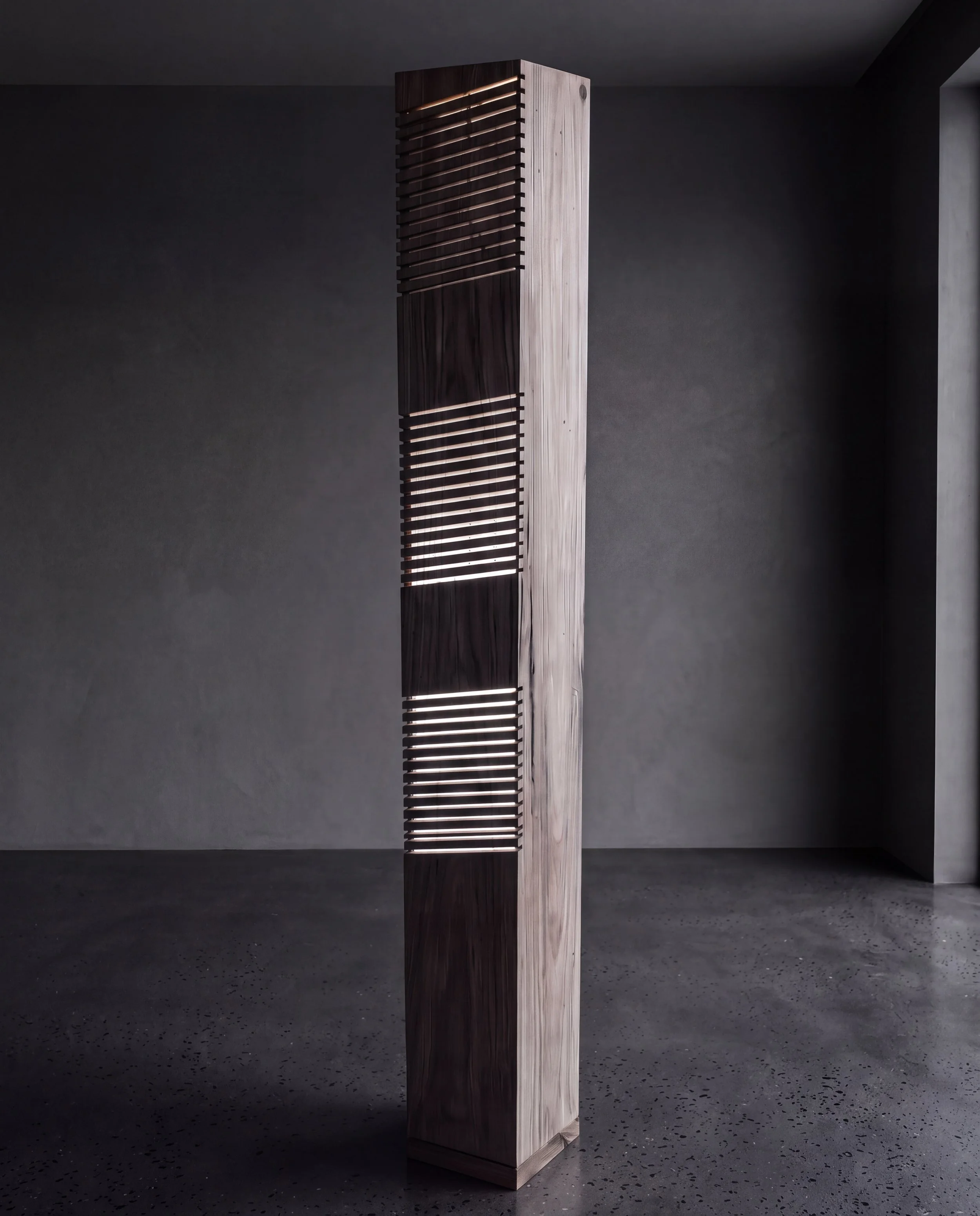 A tall, rectangular wooden sculpture with horizontal slats and a geometric design, standing on a speckled concrete floor against a dark wall.