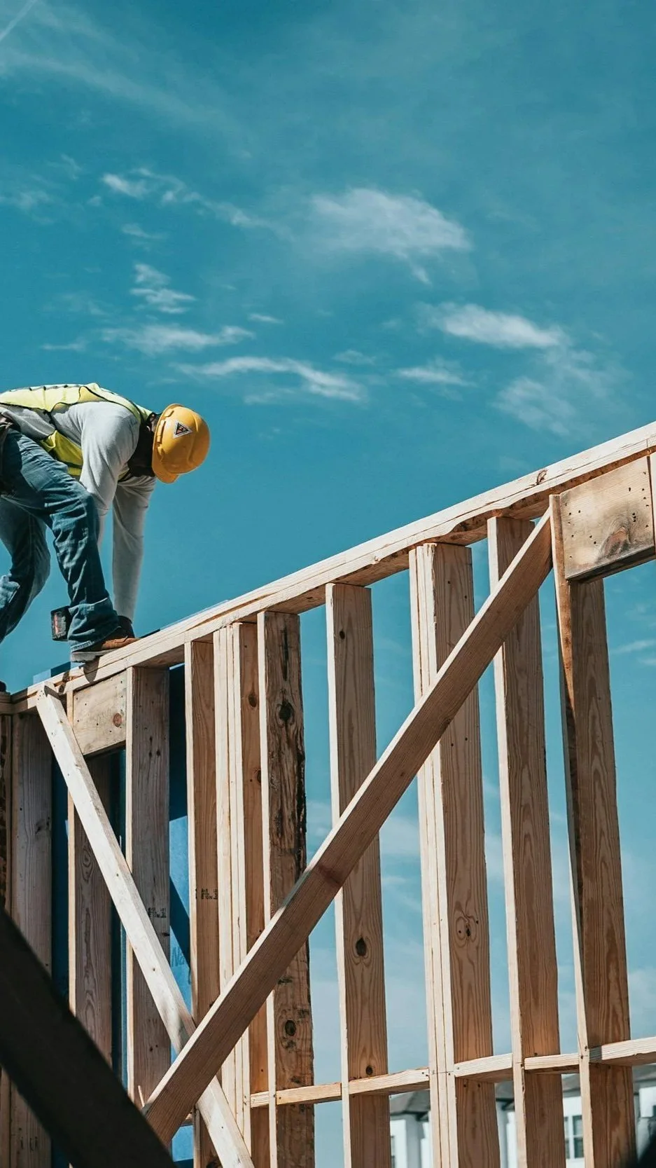 A construction worker wearing a yellow safety helmet and gray shirt is working on a wooden frame of a building under a blue sky with some clouds.