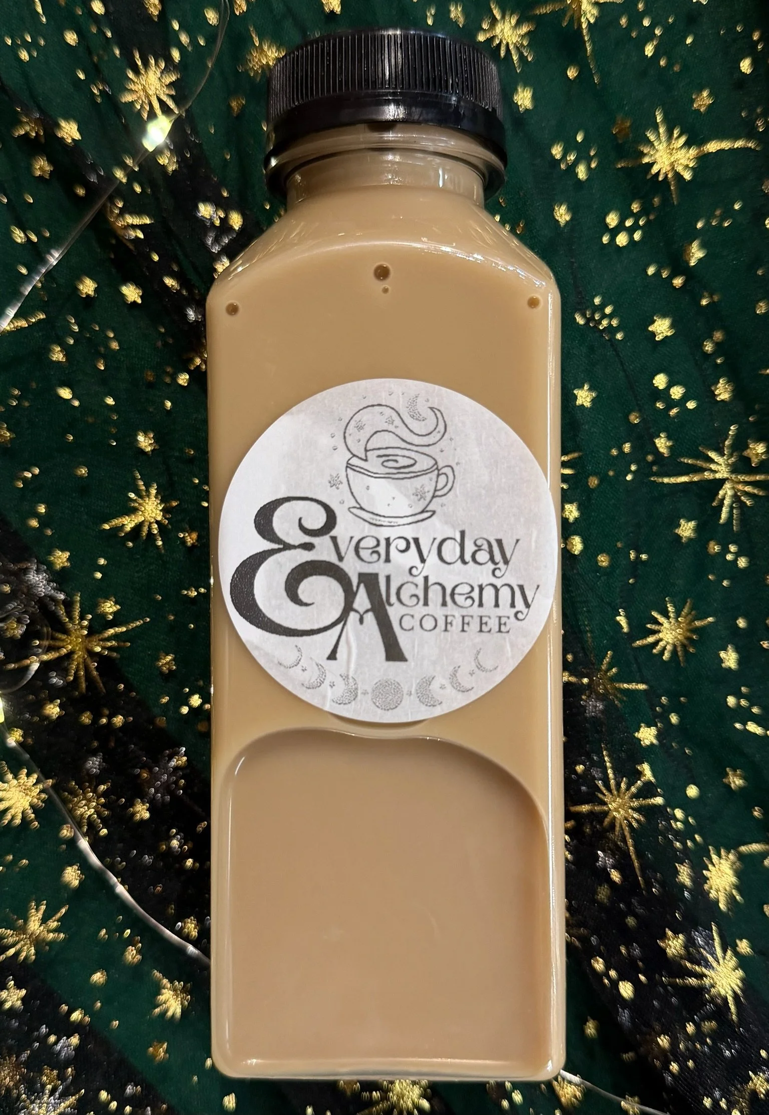 Everyday Alchemy bottled latte on dark green fabric patterned with gold stars and moons.