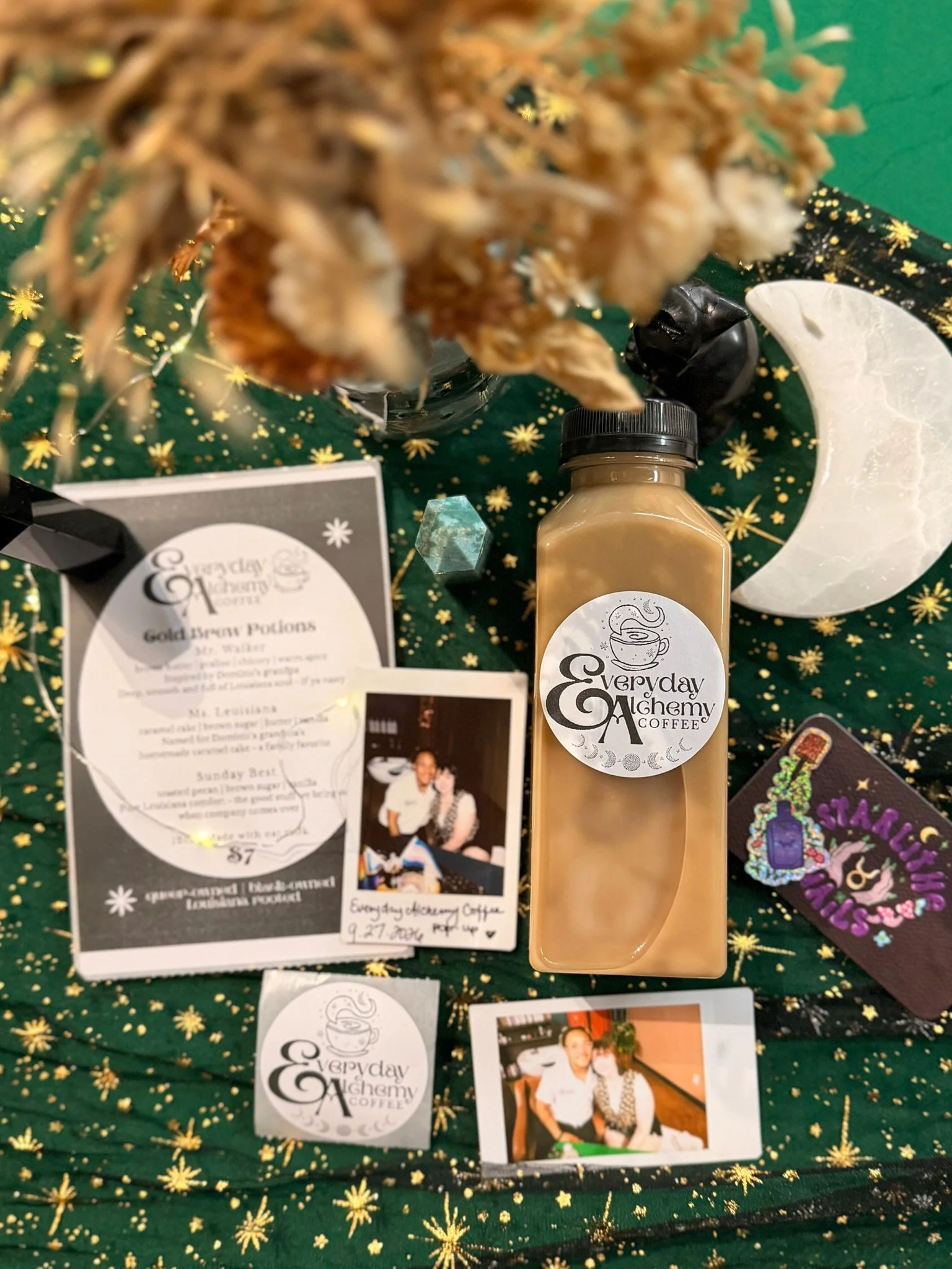 Everyday Alchemy Coffee display with a bottled latte, glass jars, a crescent moon decoration and printed cards on green fabric with gold stars.