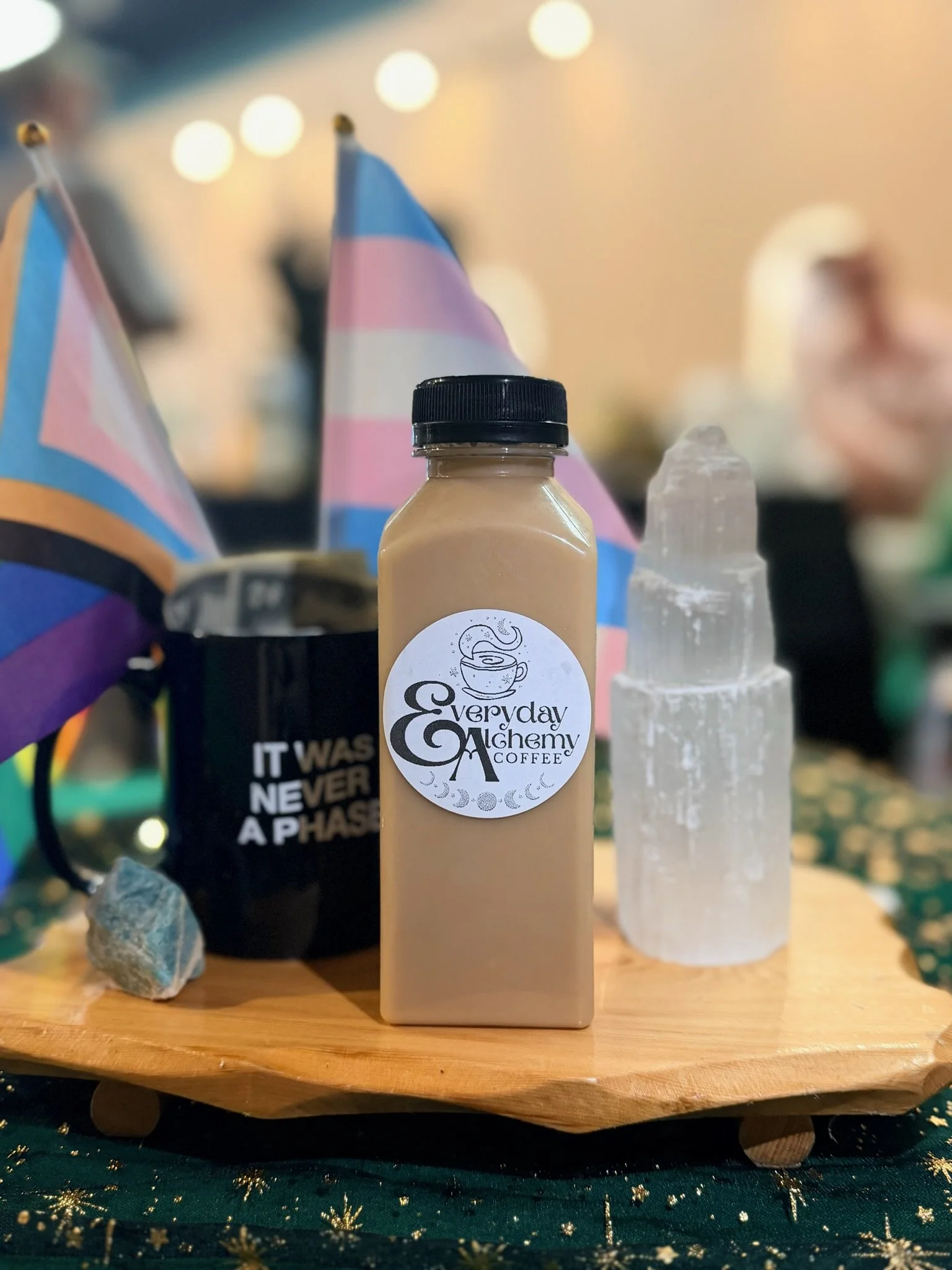 Everyday Alchemy bottled latte beside Pride flags, a black mug and crystals.