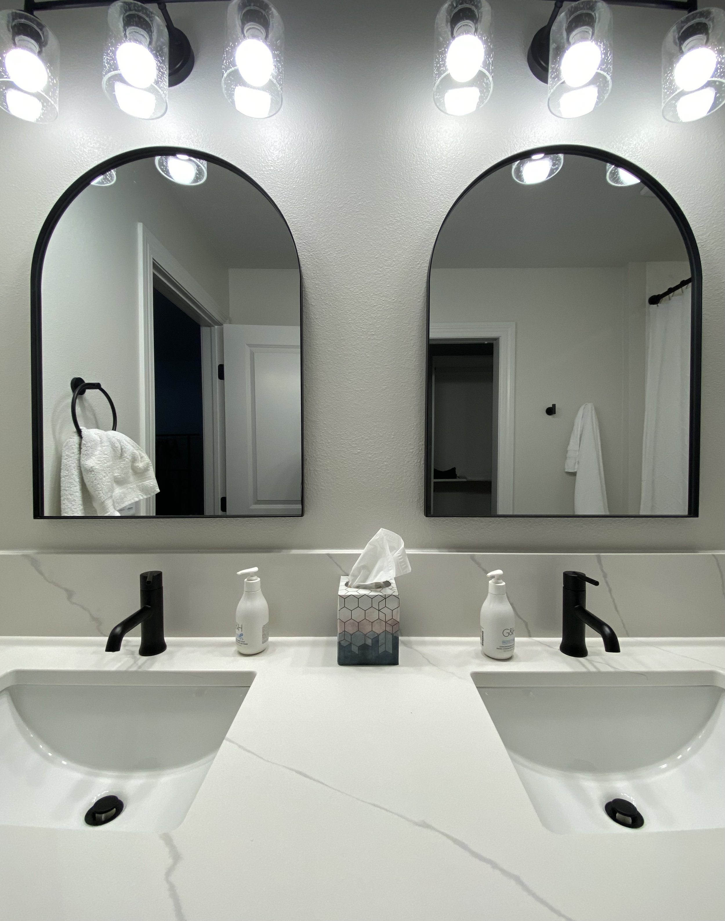 Bathroom vanity with two white sinks, black fixtures, two mirrors, and a marble countertop. Items on the counter include tissue box and hand soap. Towel is hanging on the wall, and lights are above the mirrors.