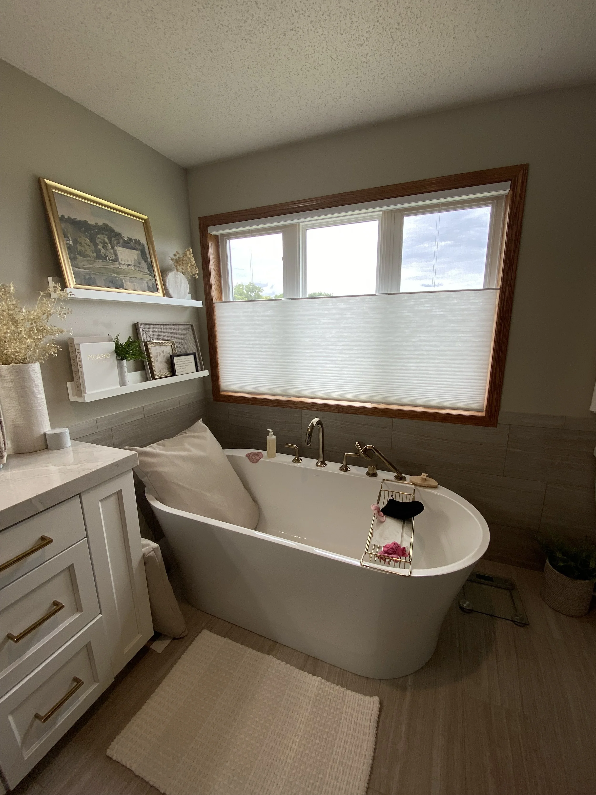 A cozy bathroom with a large window covered by a white shade, a white bathtub with brass fixtures, a white vanity with gold handles, decorative books and picture frames, a beige rug, and framed artwork and vases on a wall shelf.