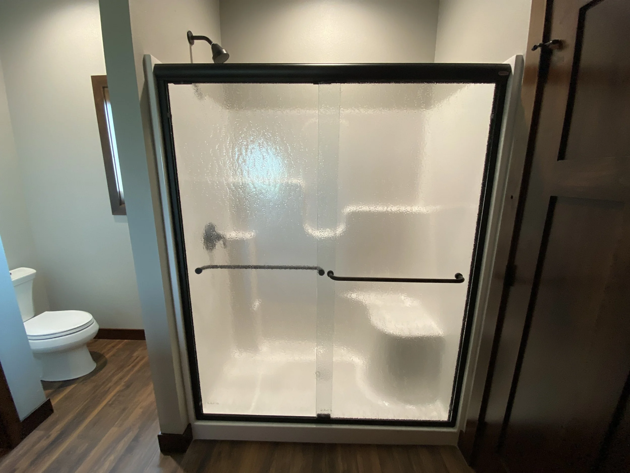 A glass shower door with black framing and a black grab bar on the inside, with a frosted lower half, in a bathroom with wooden flooring and a toilet visible to the left