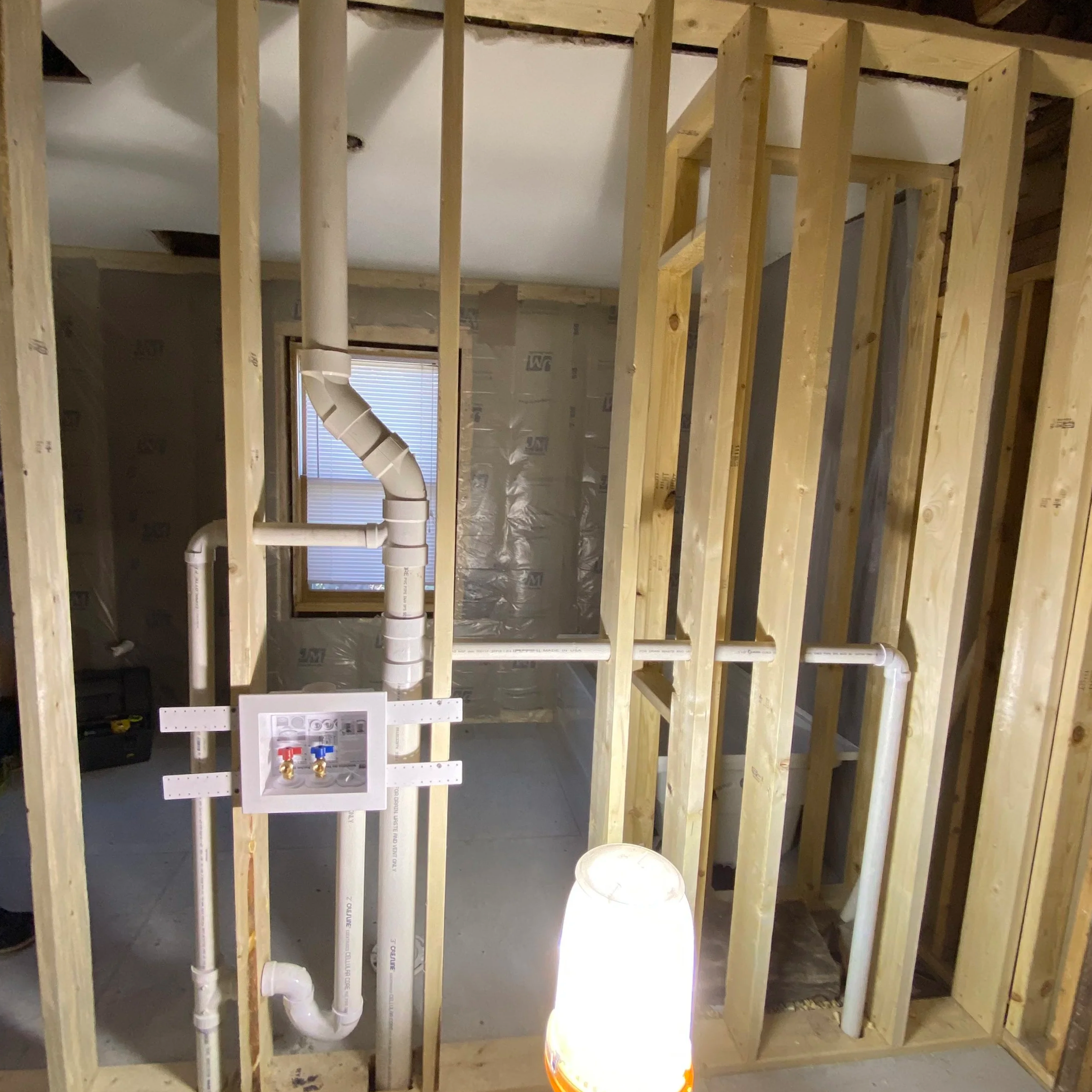 Wooden framing of a wall under construction with visible white PVC plumbing pipes, a window, and insulation in the background.