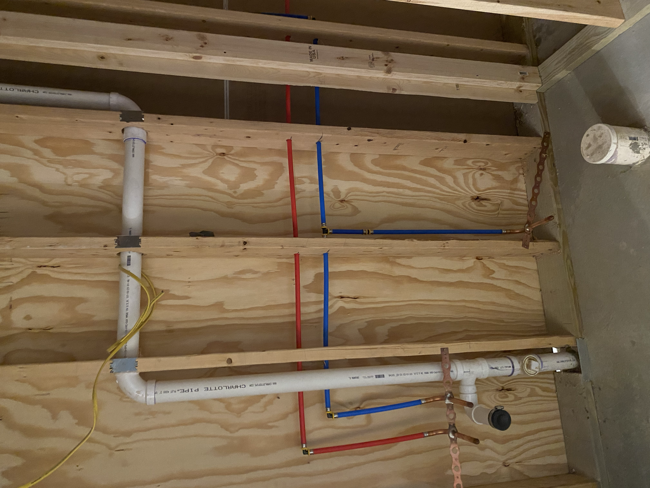 Underside view of a partially constructed wall with plumbing pipes in a building renovation. Visible pipes include white PVC, copper, and red and blue PEX tubing, mounted on wooden framing.
