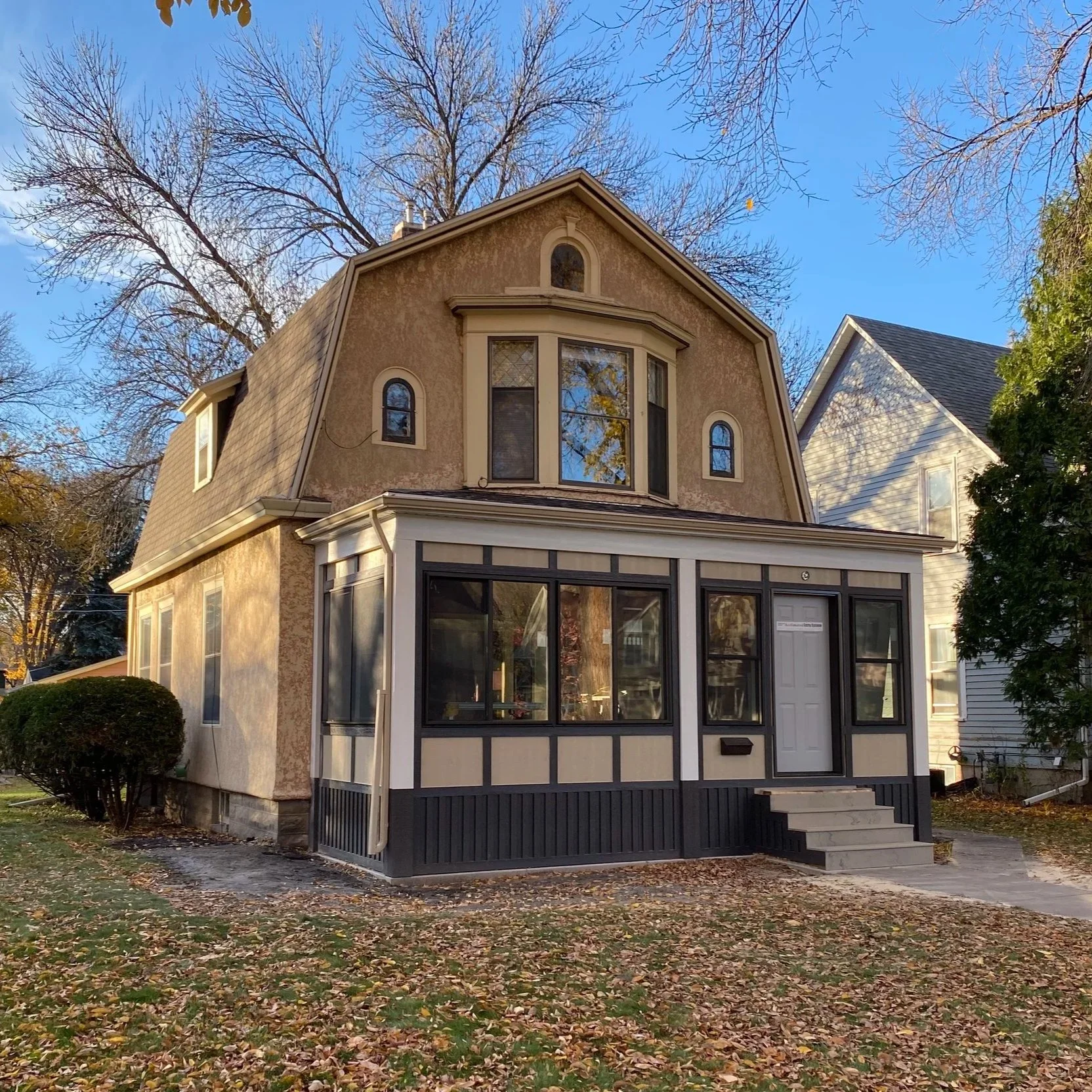 A two-story house with multiple windows, a front porch, and a sunroom with large glass panels, set against a fall landscape with trees and fallen leaves.