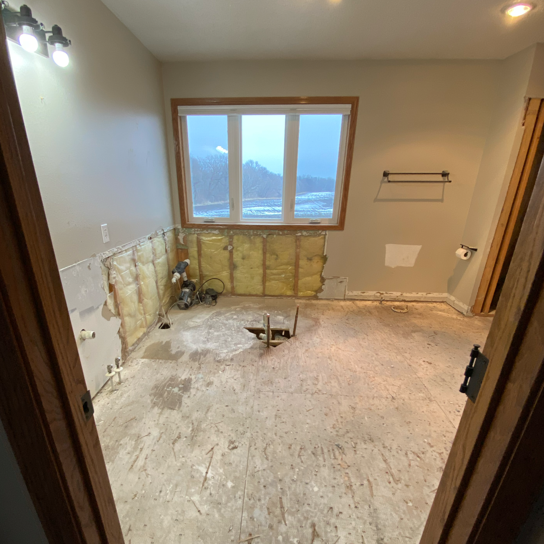 Room undergoing renovation with exposed insulation, plumbing pipes, and incomplete flooring, viewed through a doorway. A large window shows a winter landscape outside.