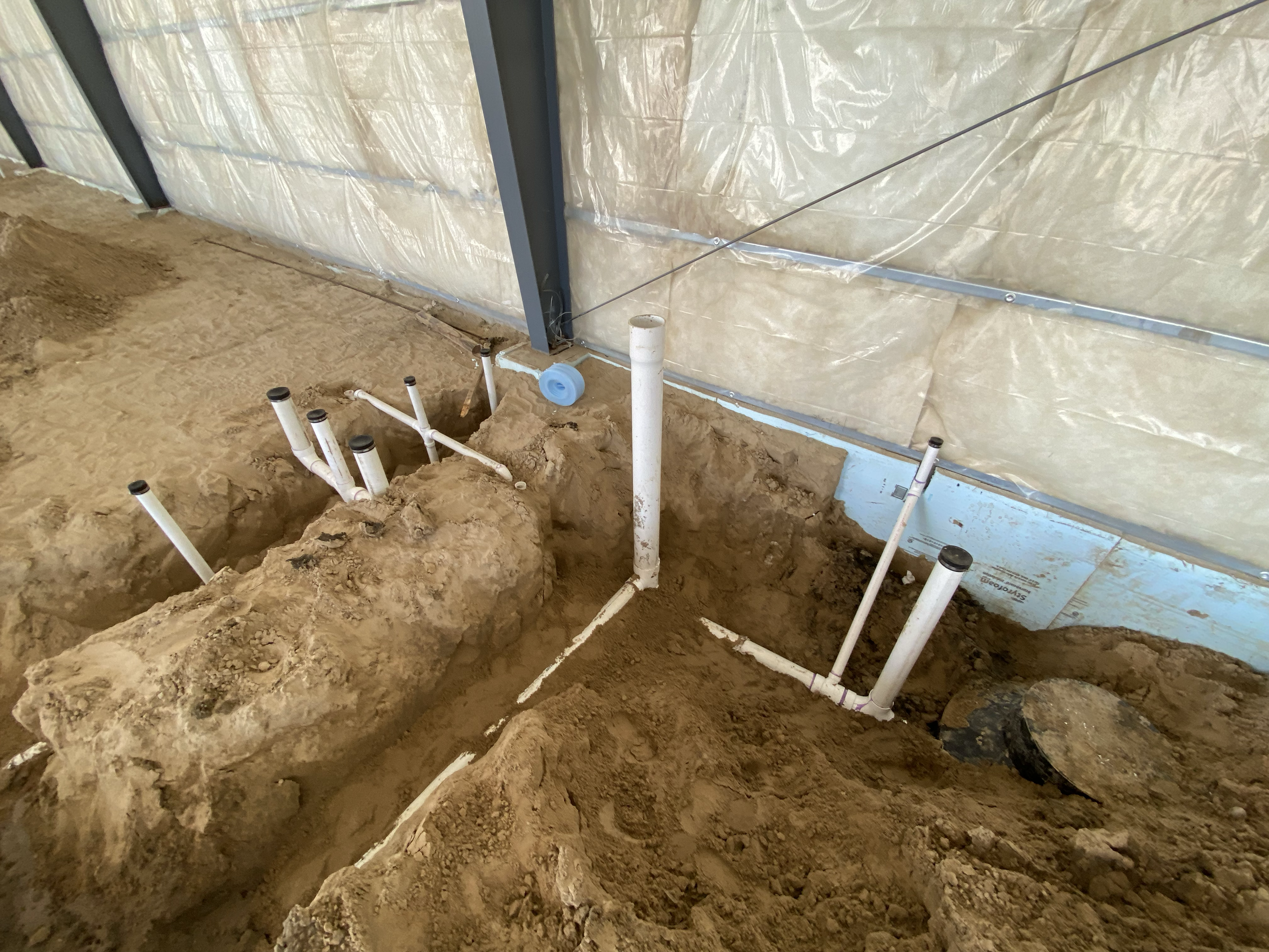Underground plumbing installation with white PVC pipes and a large white pipe elbow, sandy soil, and a plastic wall with metal framing and plastic sheeting.