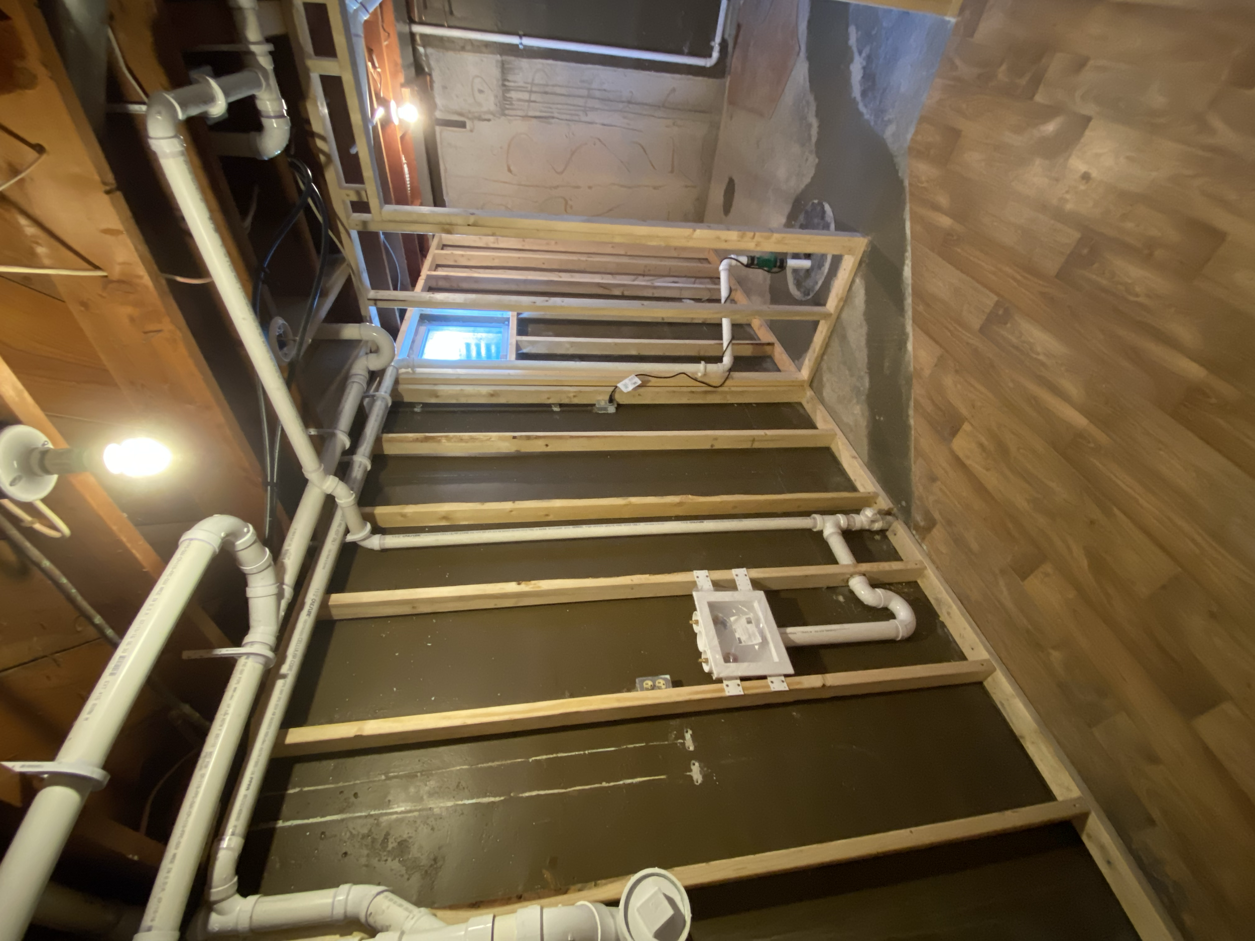 Construction site with exposed wooden framing, white PVC pipes for plumbing, and a small window in the background. The floor is partially finished with a mix of concrete and wood.