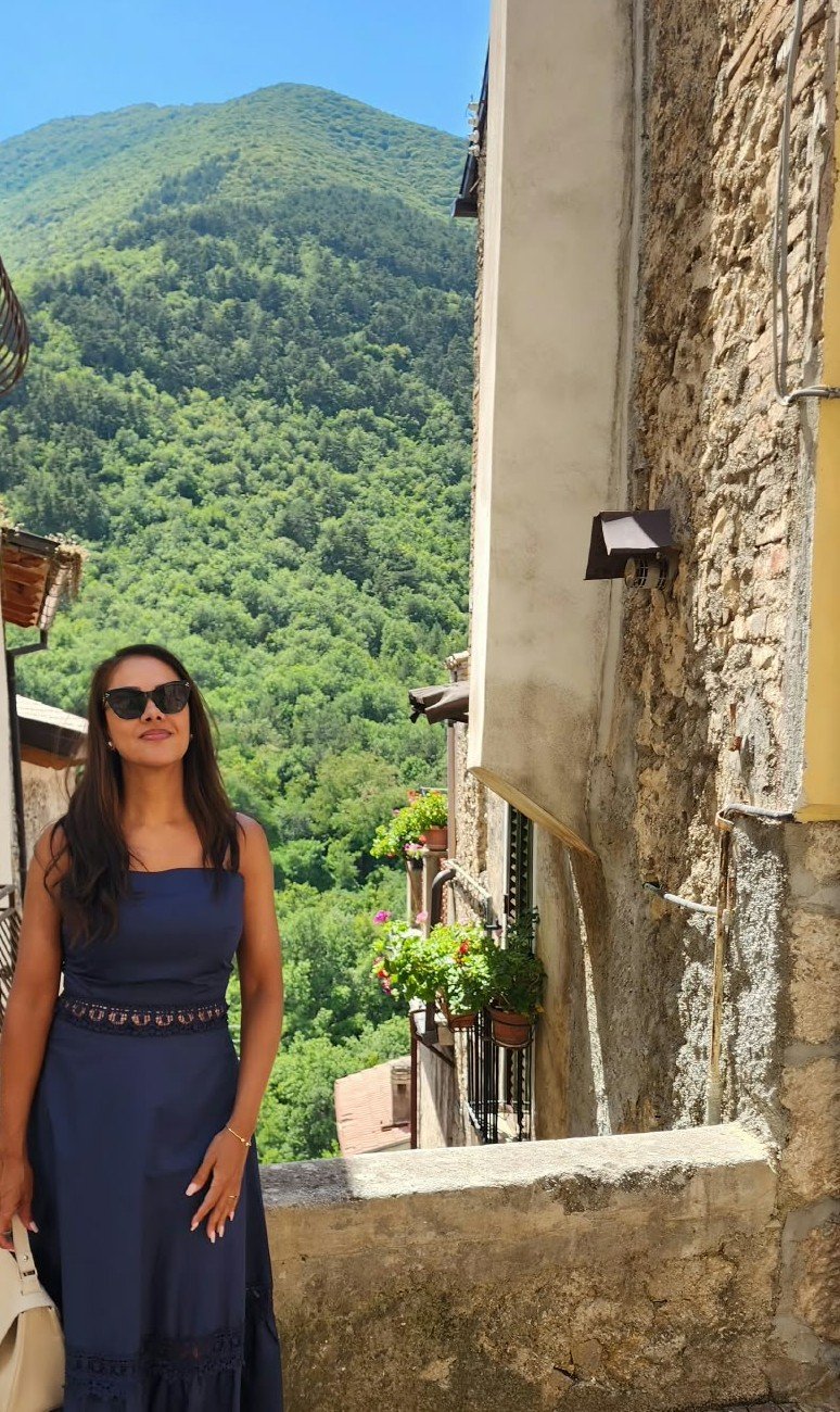Woman in a navy blue dress and sunglasses standing on a stone balcony with green hills and trees in the background. Travel to Italy and touring around a small village.