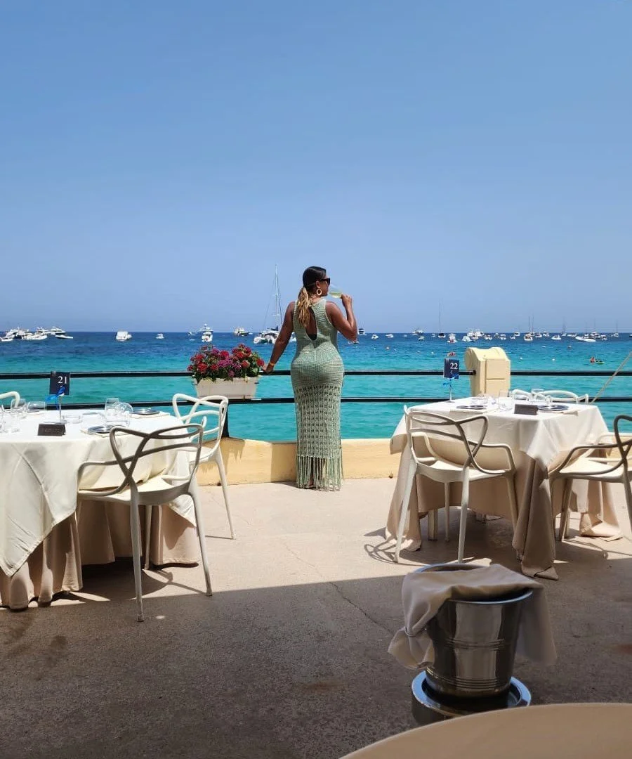 A woman in a long green crochet dress is standing on an outdoor seaside restaurant terrace, drinking from a glass, with tables and chairs around and boats in the ocean background on a clear day. A fine dining at a seaside elegant restaurant.