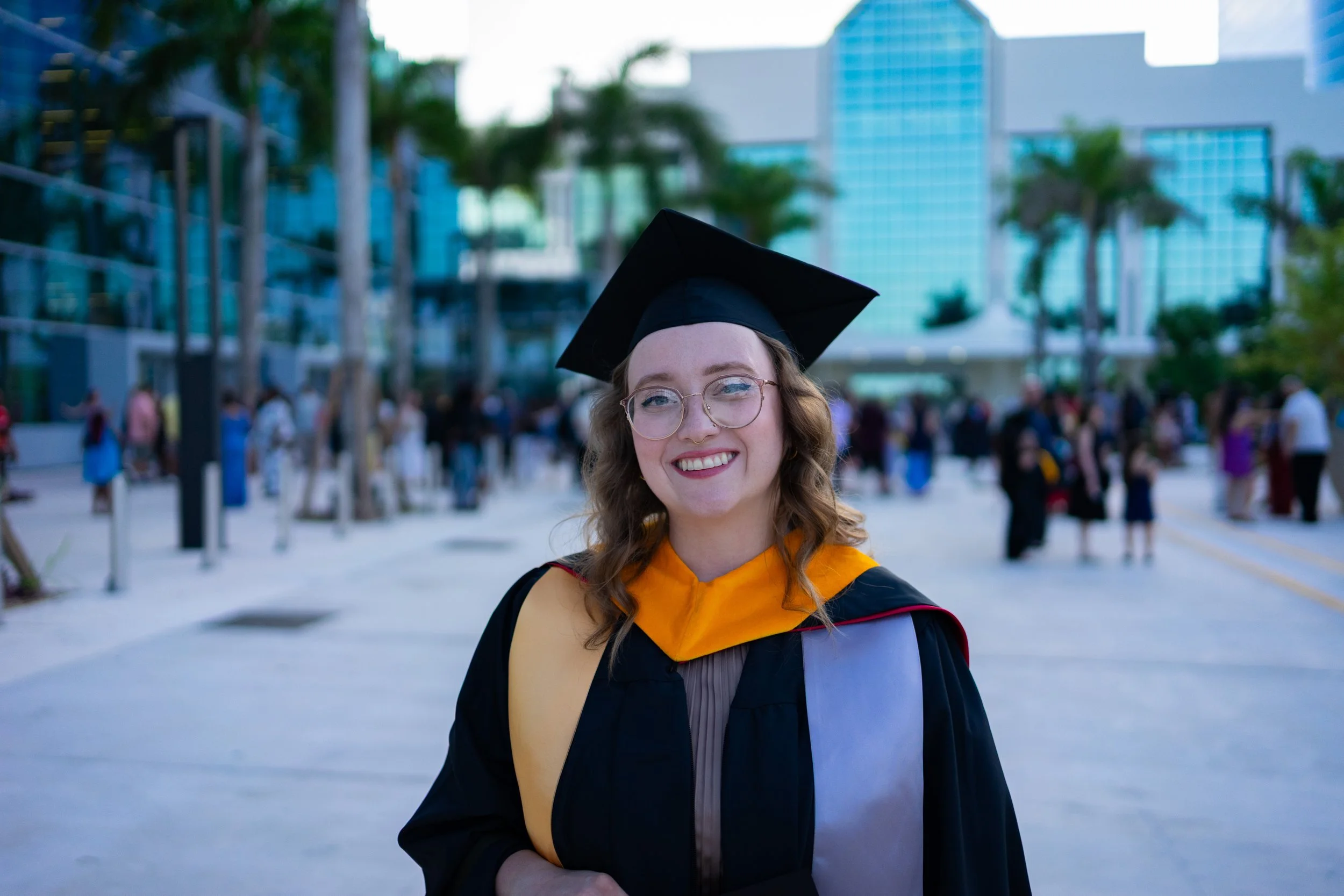 Young woman in a graduation cap and gown smiling outdoors at her graduation ceremony, with a crowd and modern buildings in the background.