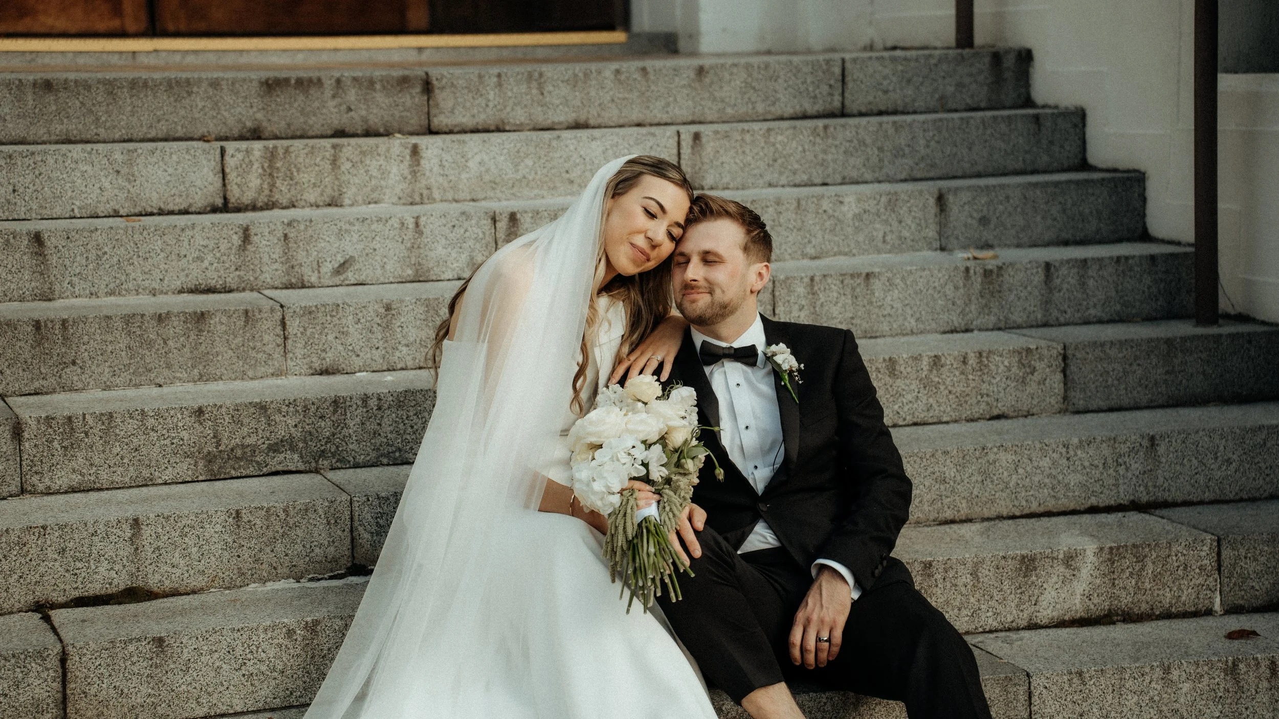 A bride and groom sitting on stone steps, leaning their heads against each other, with the bride holding a bouquet of white flowers. The bride is wearing a white wedding dress and veil, and the groom is dressed in a black tuxedo with a bow tie.