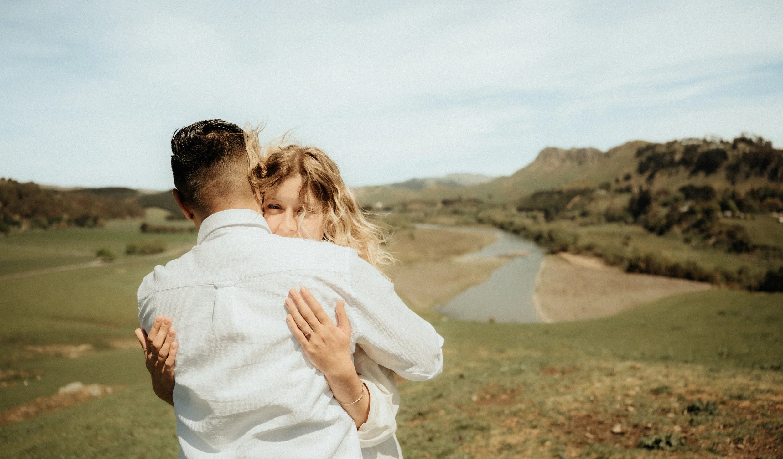 A man and woman embrace outdoors with a scenic landscape of hills, a river, and blue sky in the background.