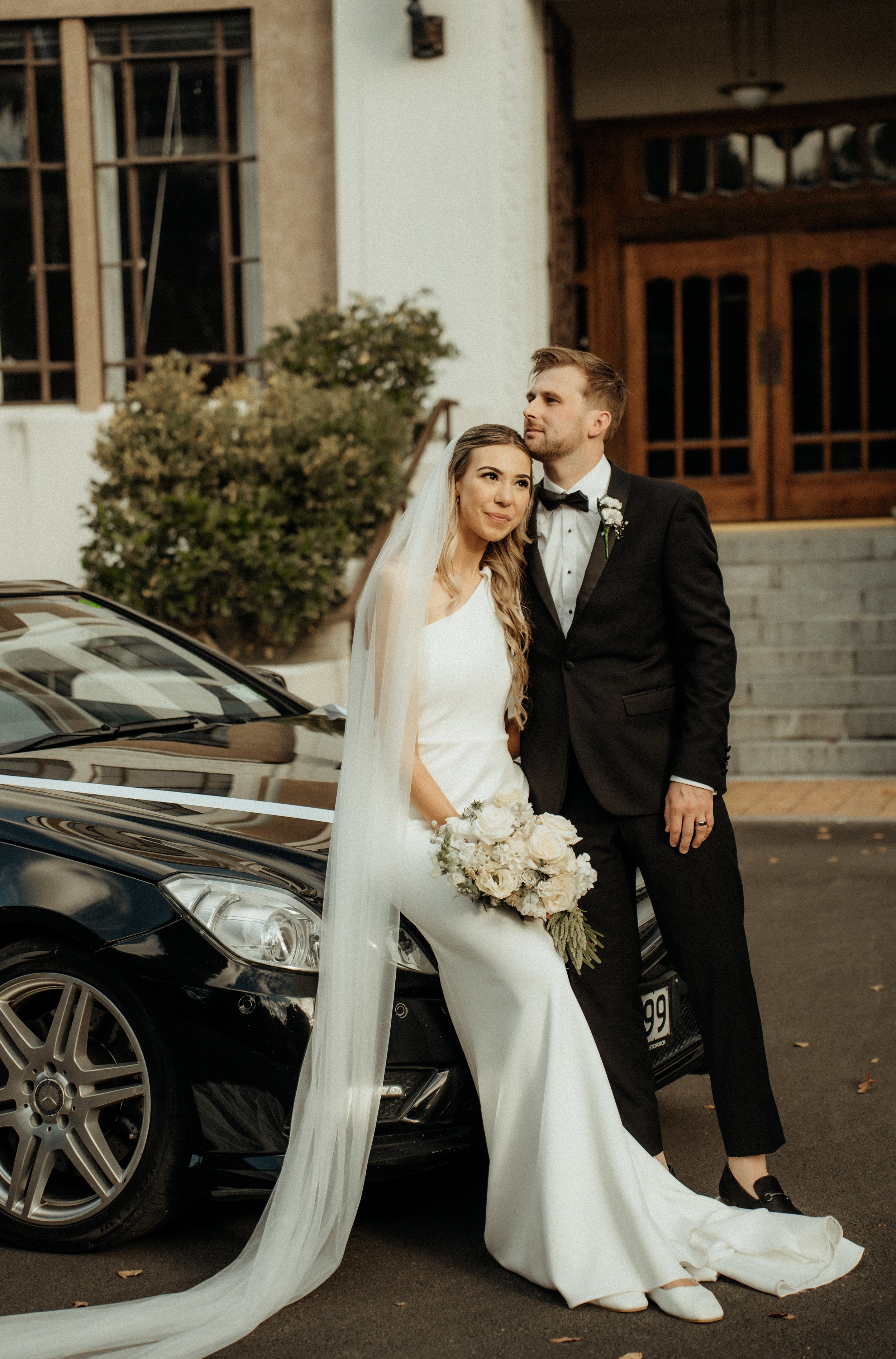 A bride and groom in wedding attire standing outside near a black luxury car, posing for a photo in front of a building with stairs and wooden doors. The bride holds a bouquet of white flowers, and the groom is dressed in a black tuxedo with a bow ti