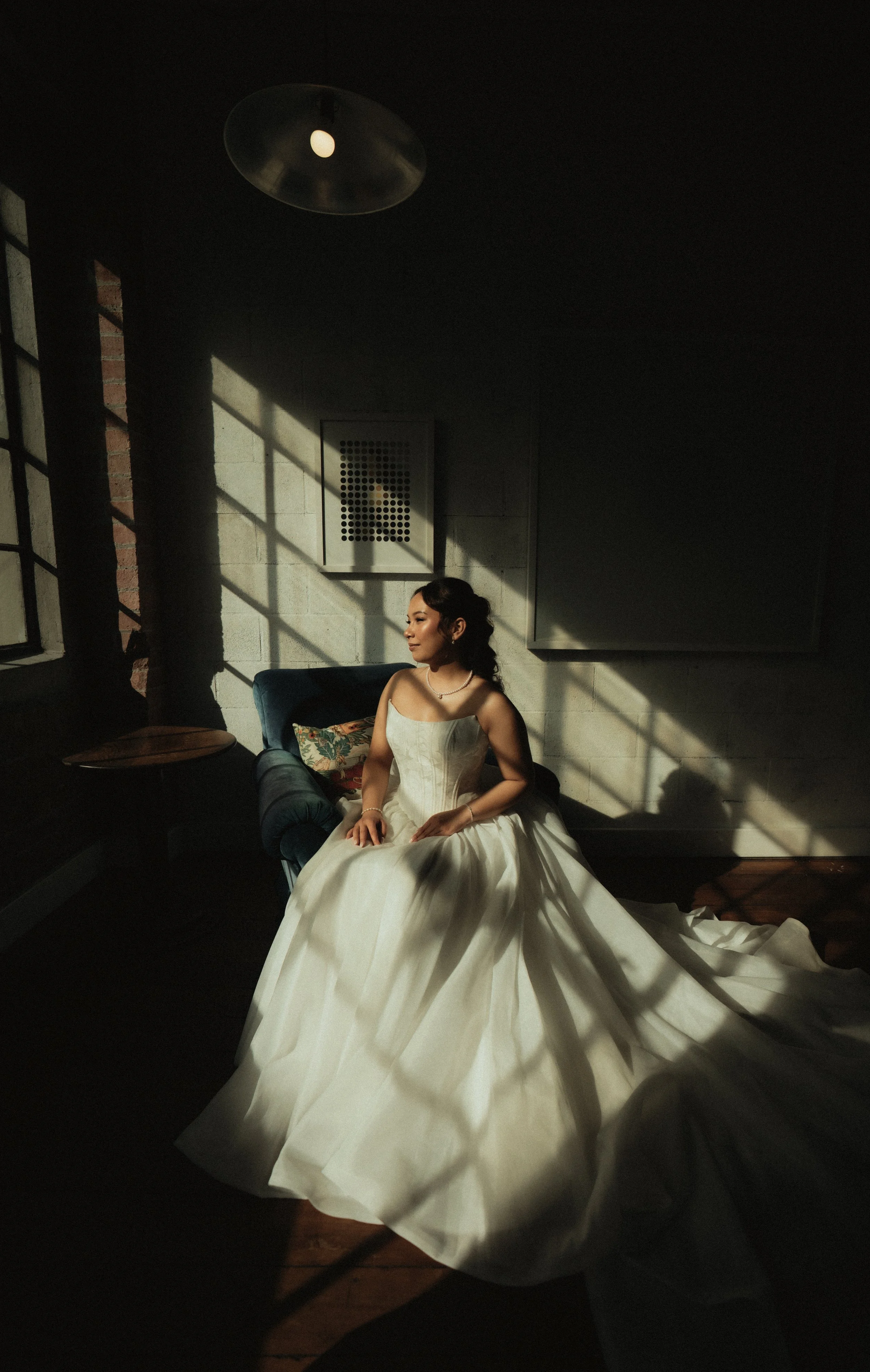 A woman in a white wedding gown sitting on a dark blue couch in a dimly lit room with sunlight streaming through a window, casting shadows on the wall behind her.