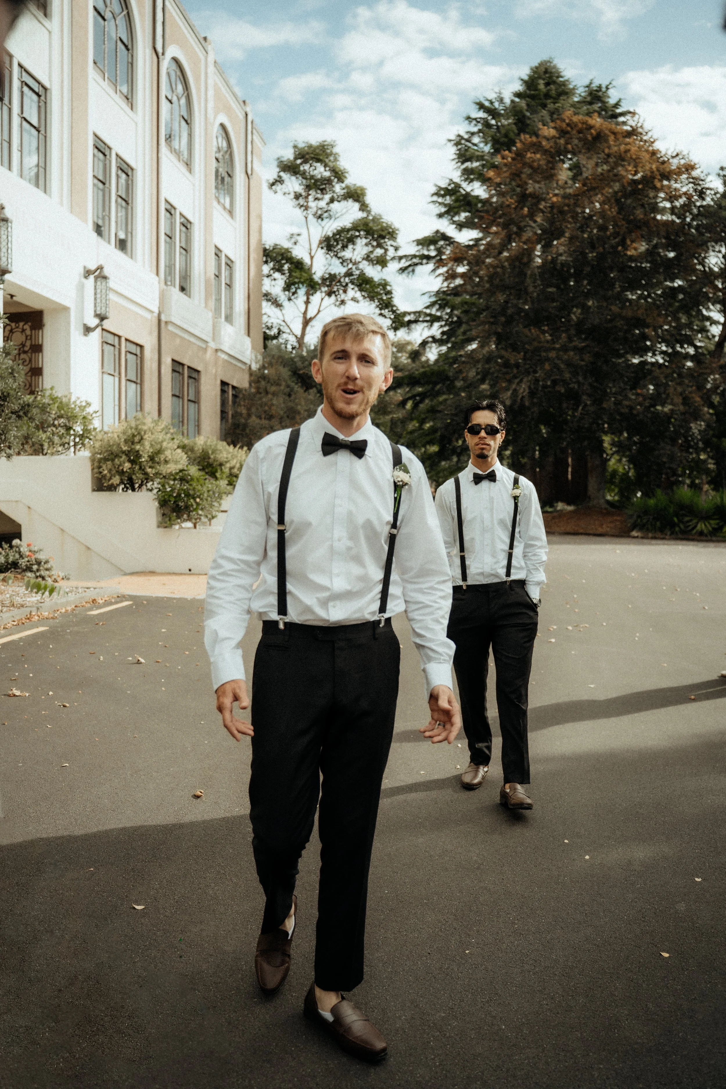Two men dressed in formal attire, including white dress shirts, black suspenders, black pants, and bow ties, walking outdoors on a paved path. The man in the foreground has blonde hair and facial hair, and is smiling with his mouth open. The man in t