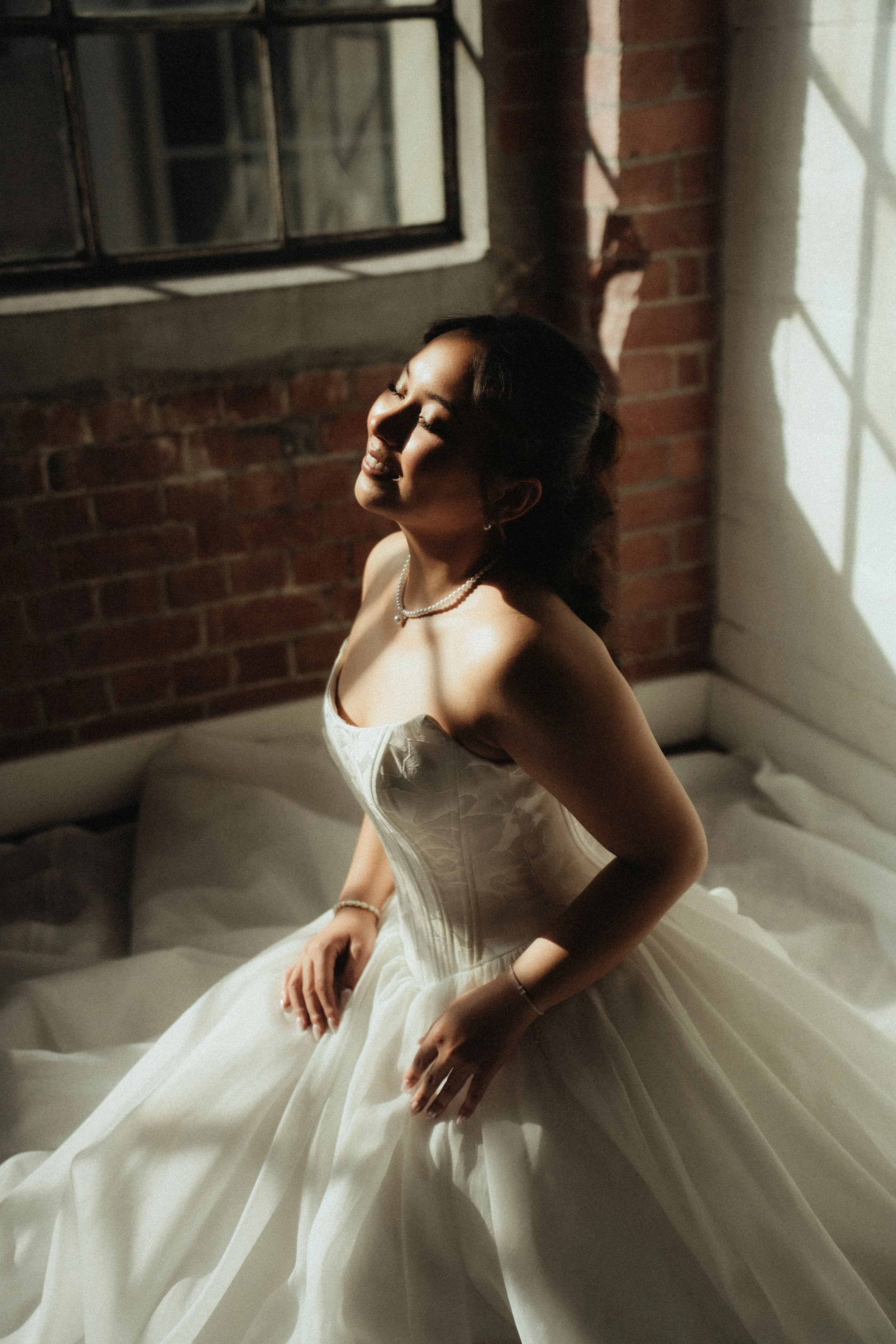 A woman in a wedding dress sitting on a bed with a brick wall and a window casting shadows behind her, smiling with closed eyes.
