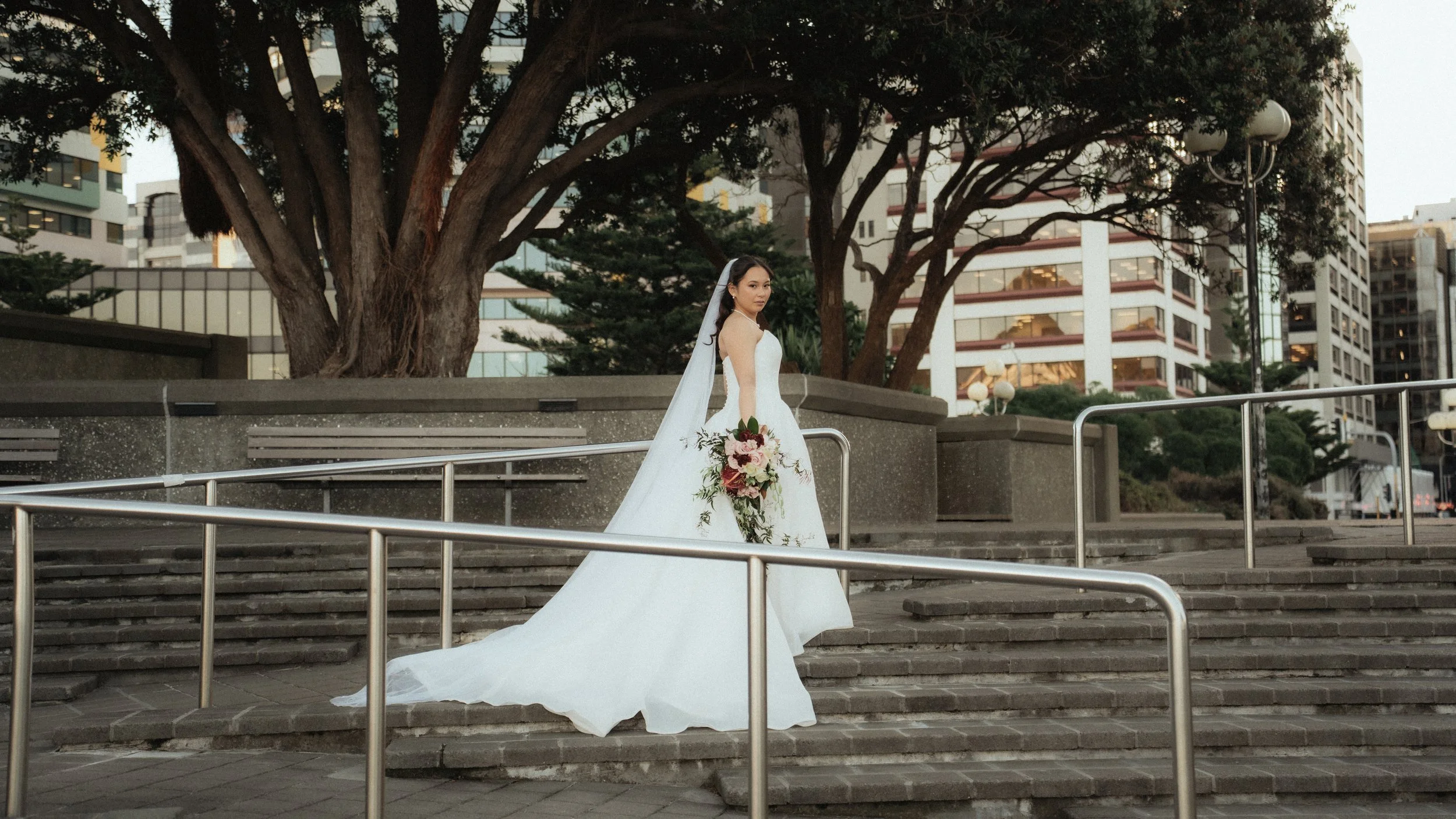A bride in a white wedding gown holding a bouquet of flowers, standing on outdoor steps with a large tree and modern buildings in the background.