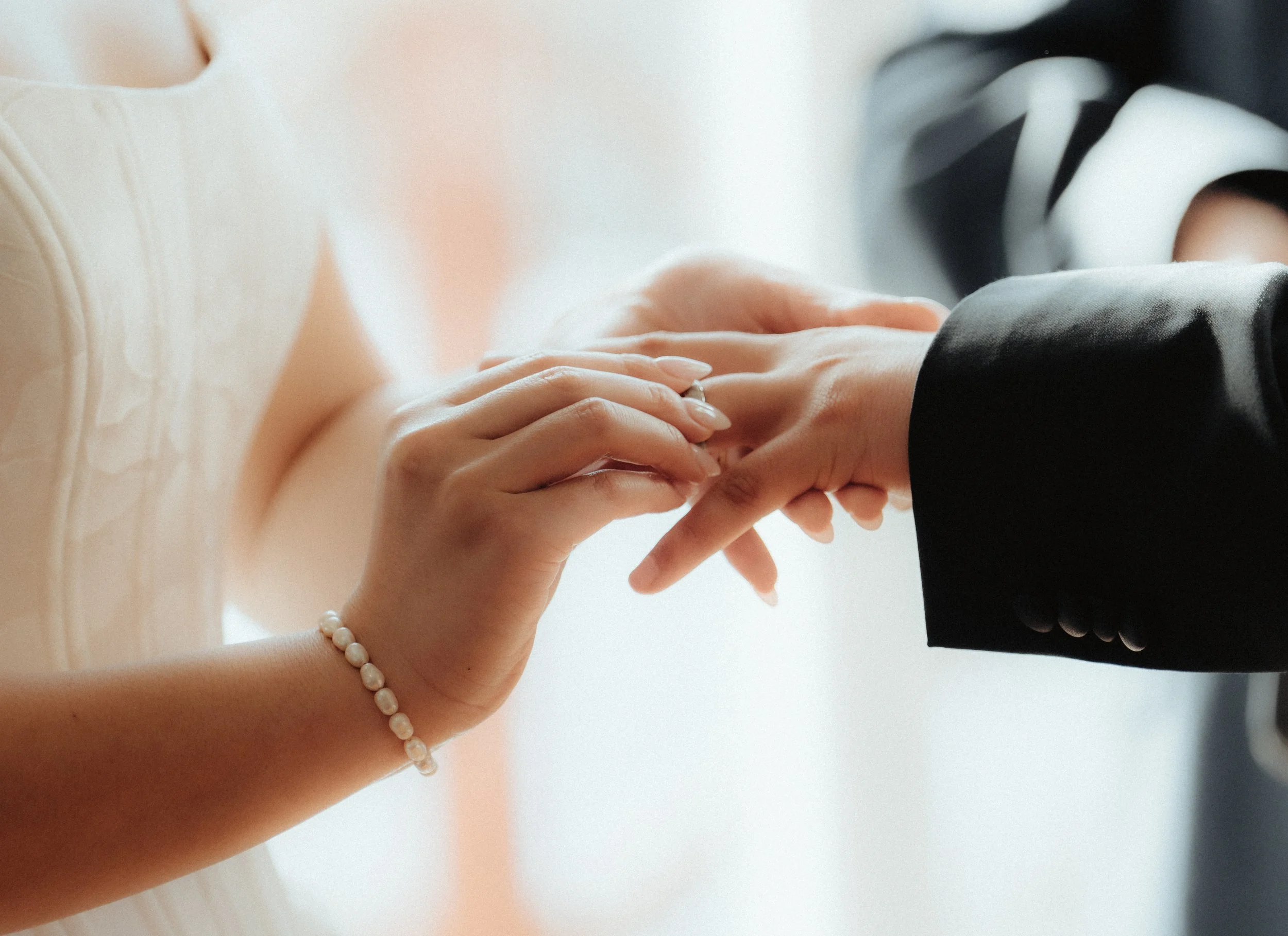 A woman wearing a pearl bracelet is holding hands with a person dressed in a black suit. Their hands are gently clasped together, indicating an intimate or romantic moment.