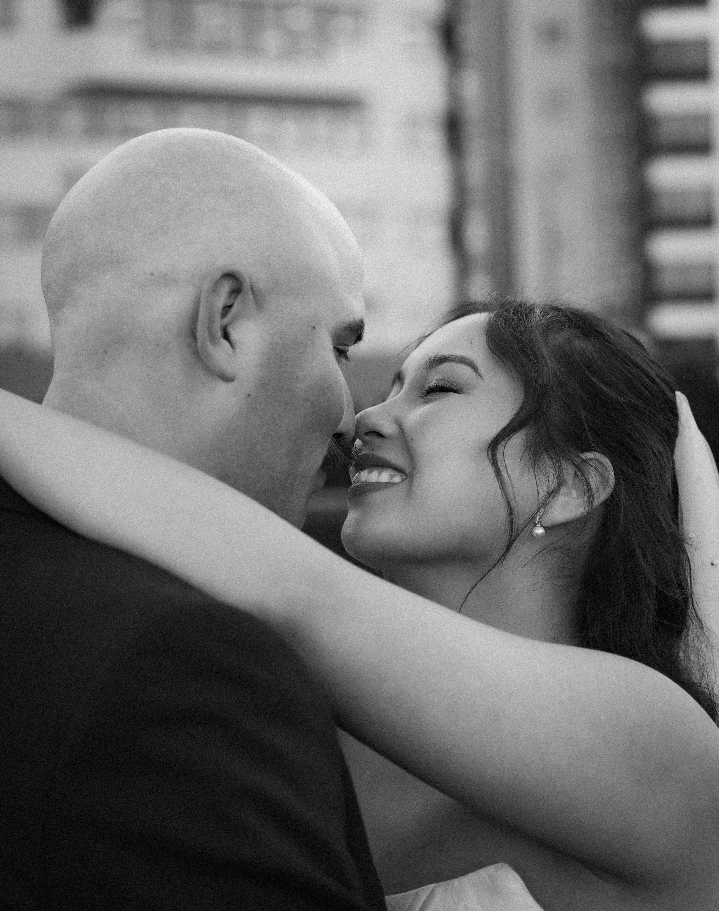 A black and white close-up photograph of a couple in an intimate moment. The man has a shaved head and is facing the woman, who has long dark hair and is smiling with closed eyes. The scene appears tender and loving.