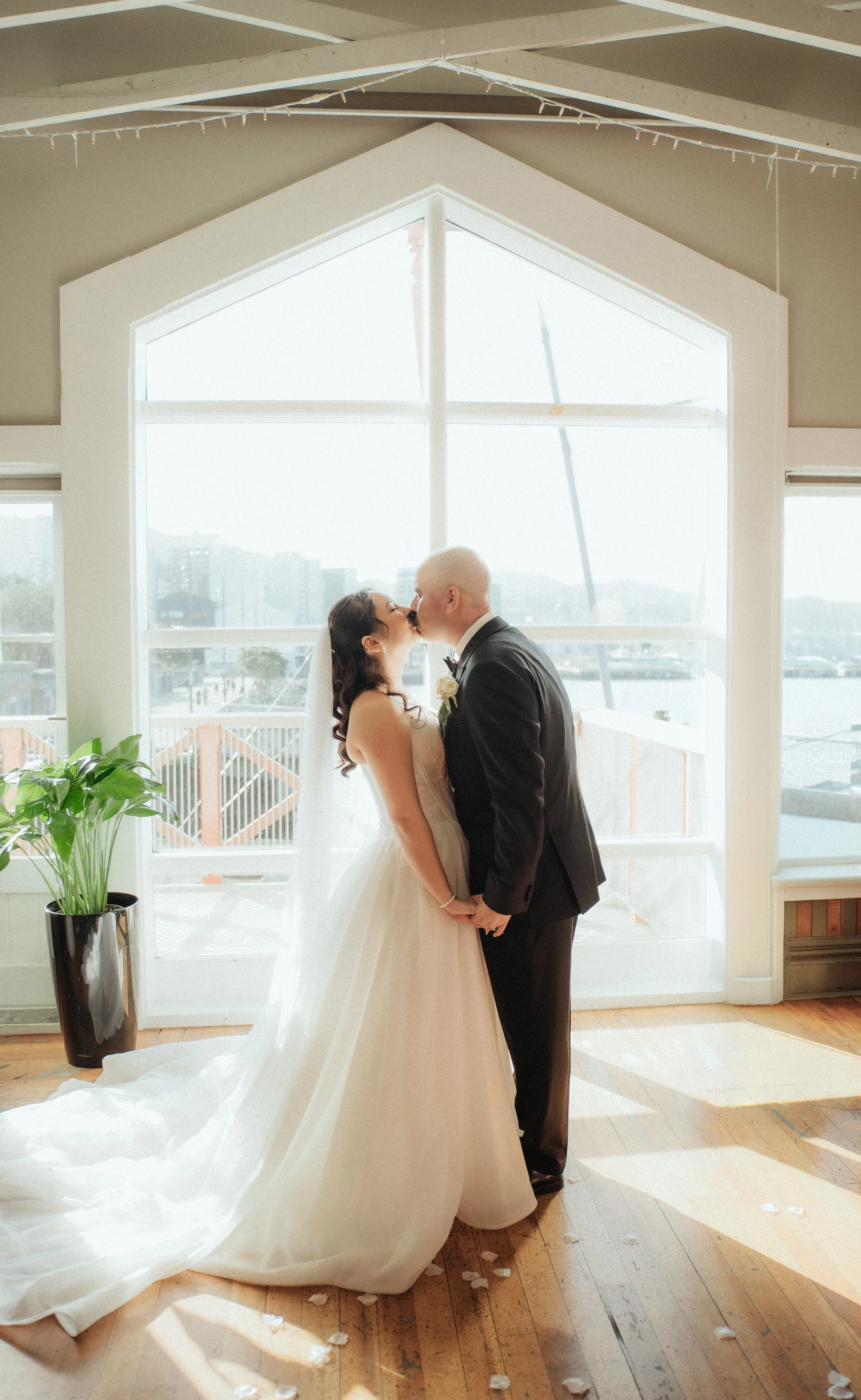A bride and groom share a kiss indoors in front of large window, holding hands, with petals on the wooden floor, a potted plant to the left, and natural daylight.