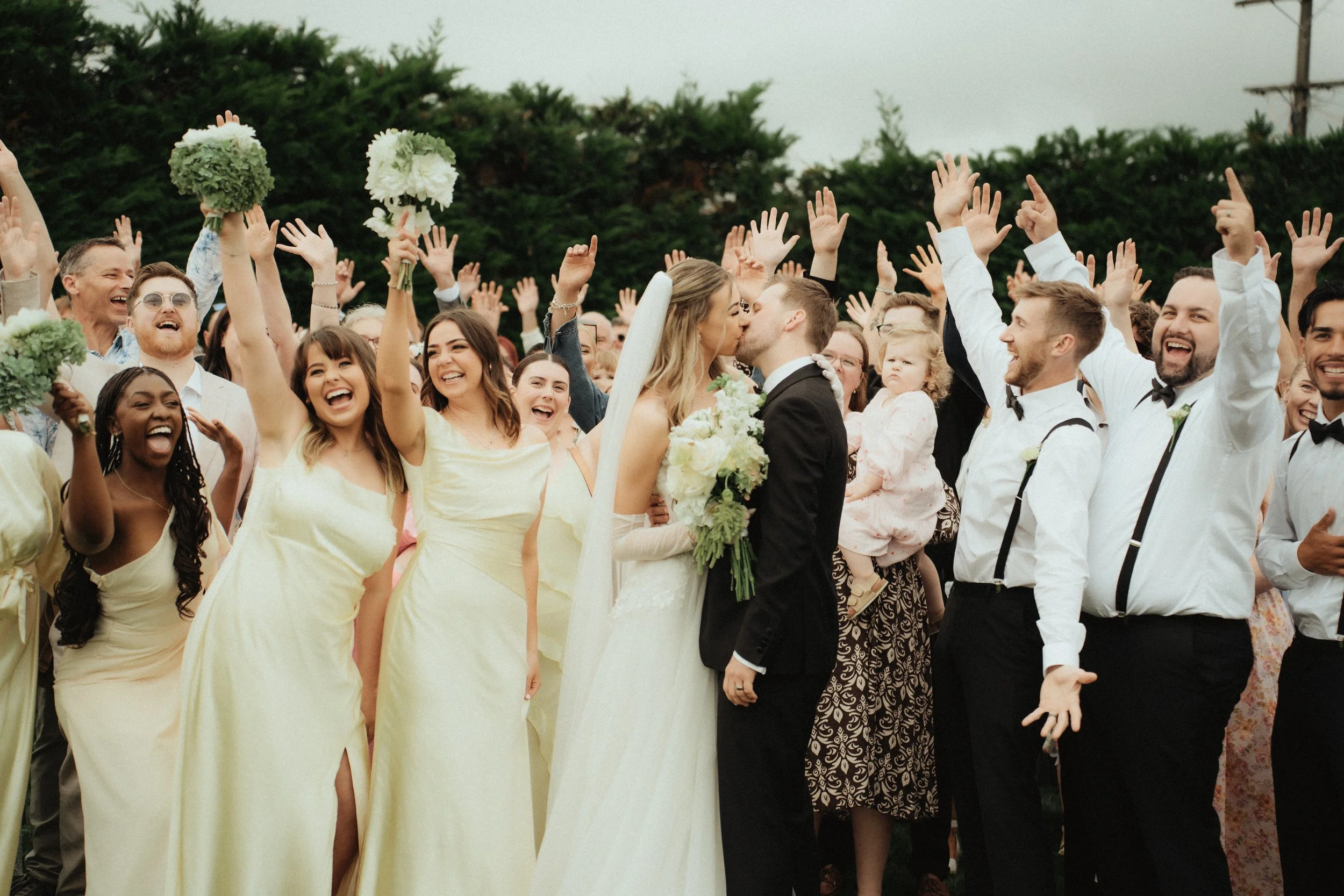 A wedding celebration with a large group of people, including a bride and groom sharing a kiss, surrounded by friends and family who are cheering and raising their hands.