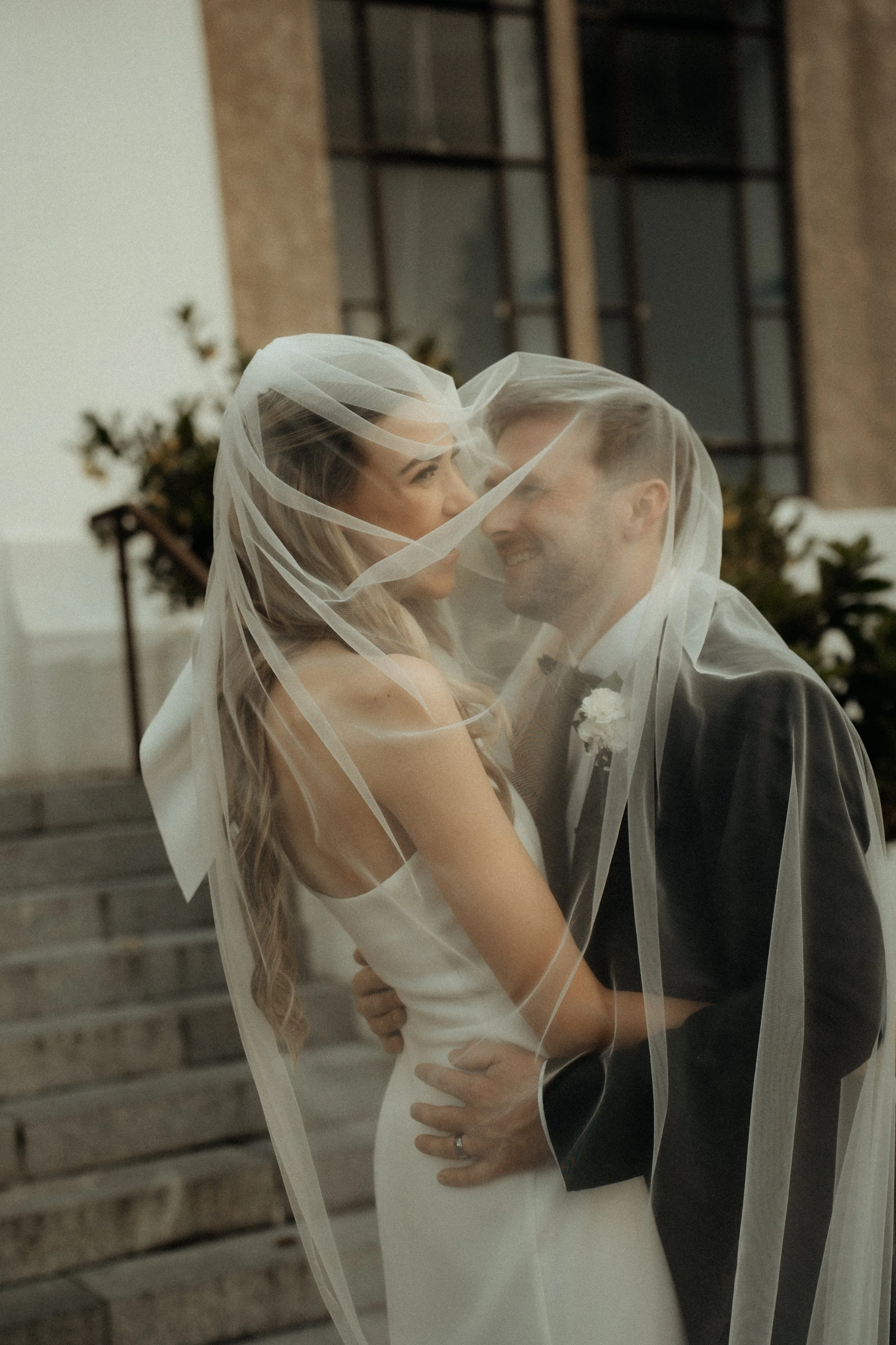 A bride and groom sharing a moment under a veil, smiling and holding each other, in an outdoor setting with brick stairs and planters in the background.