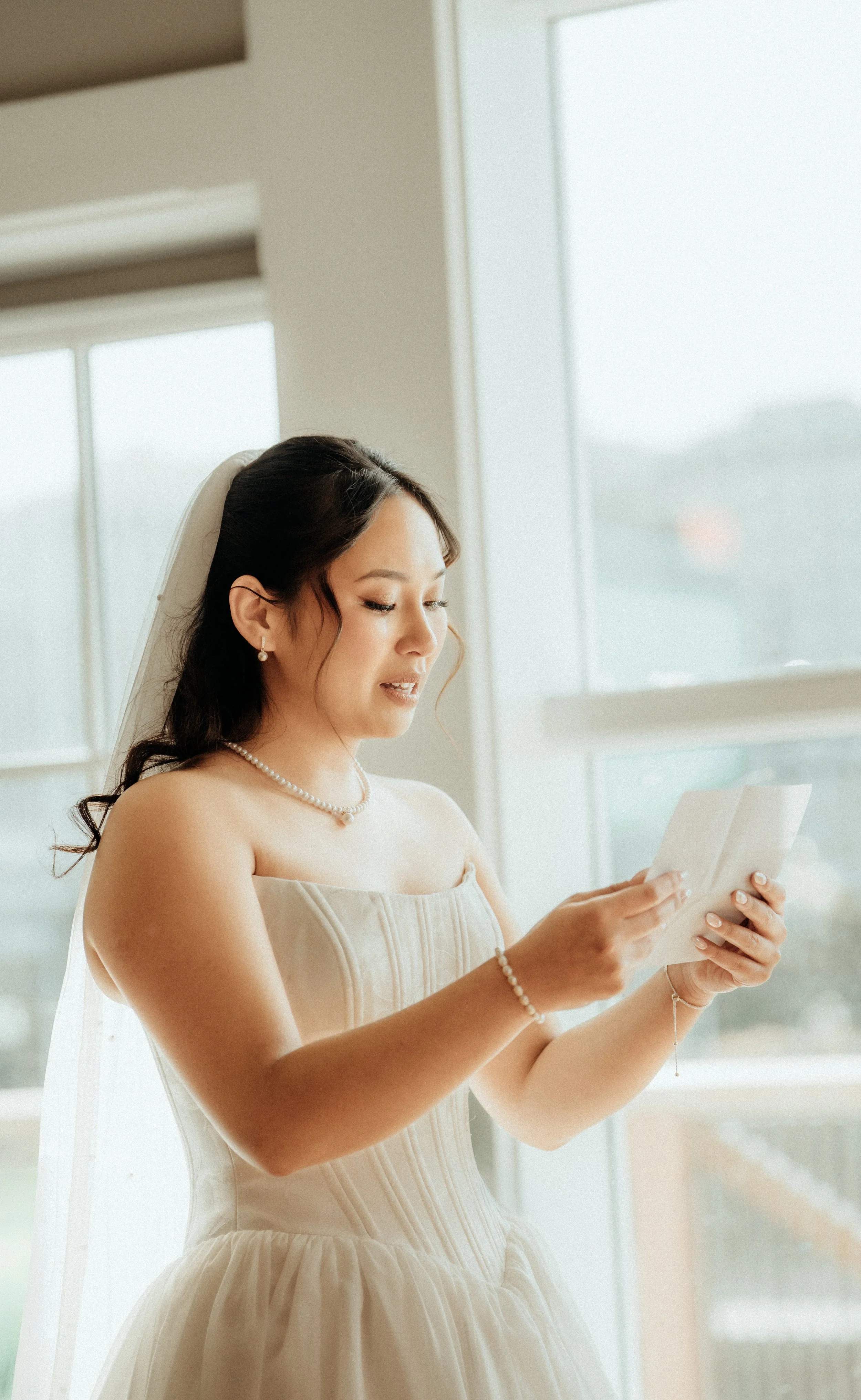 A bride in a strapless dress and pearl jewelry reads a letter or note in front of a window with natural light.