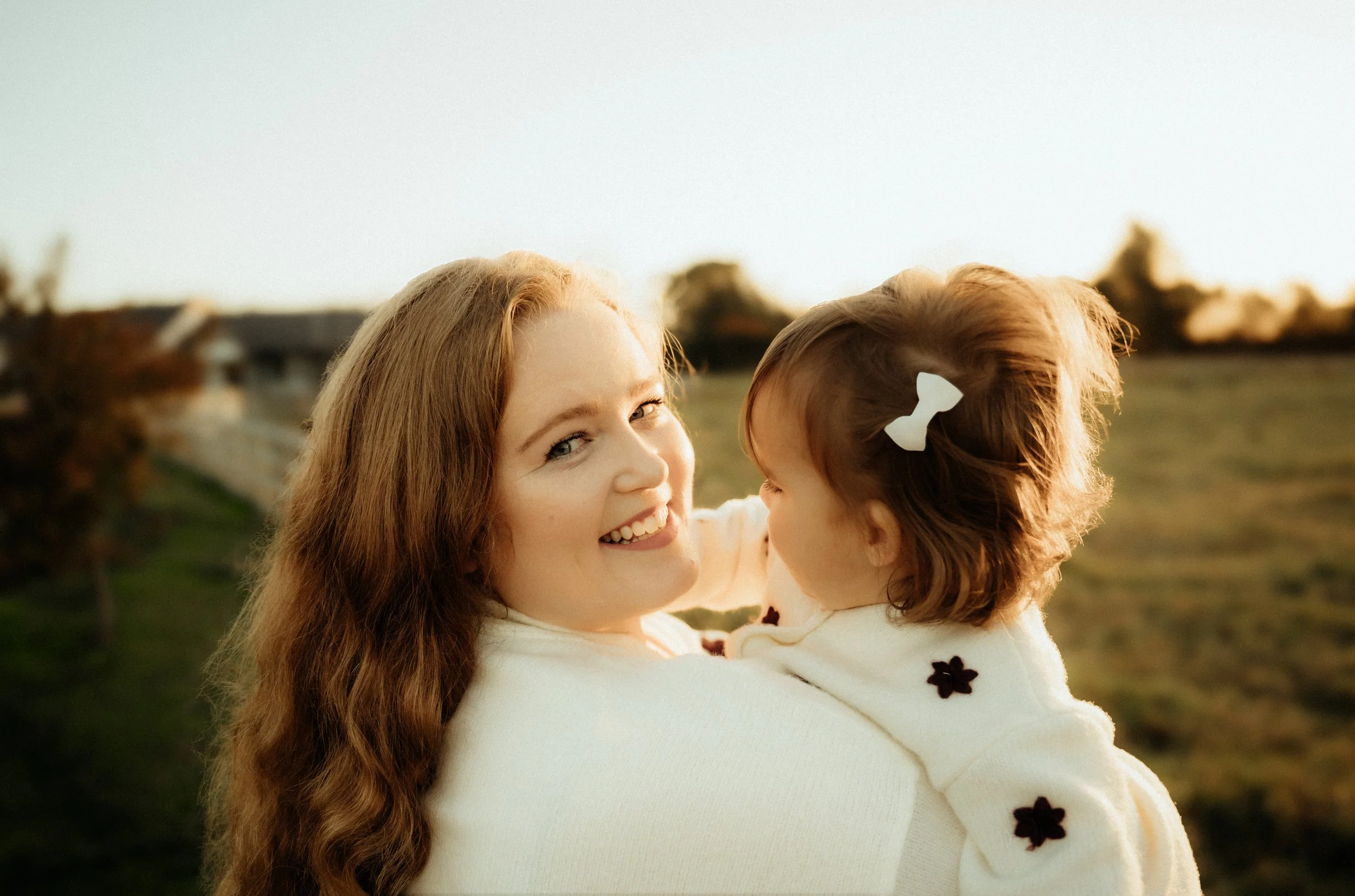 A woman with long red hair holding a young girl with brown hair and a white bow, outdoors in the late afternoon sunlight.