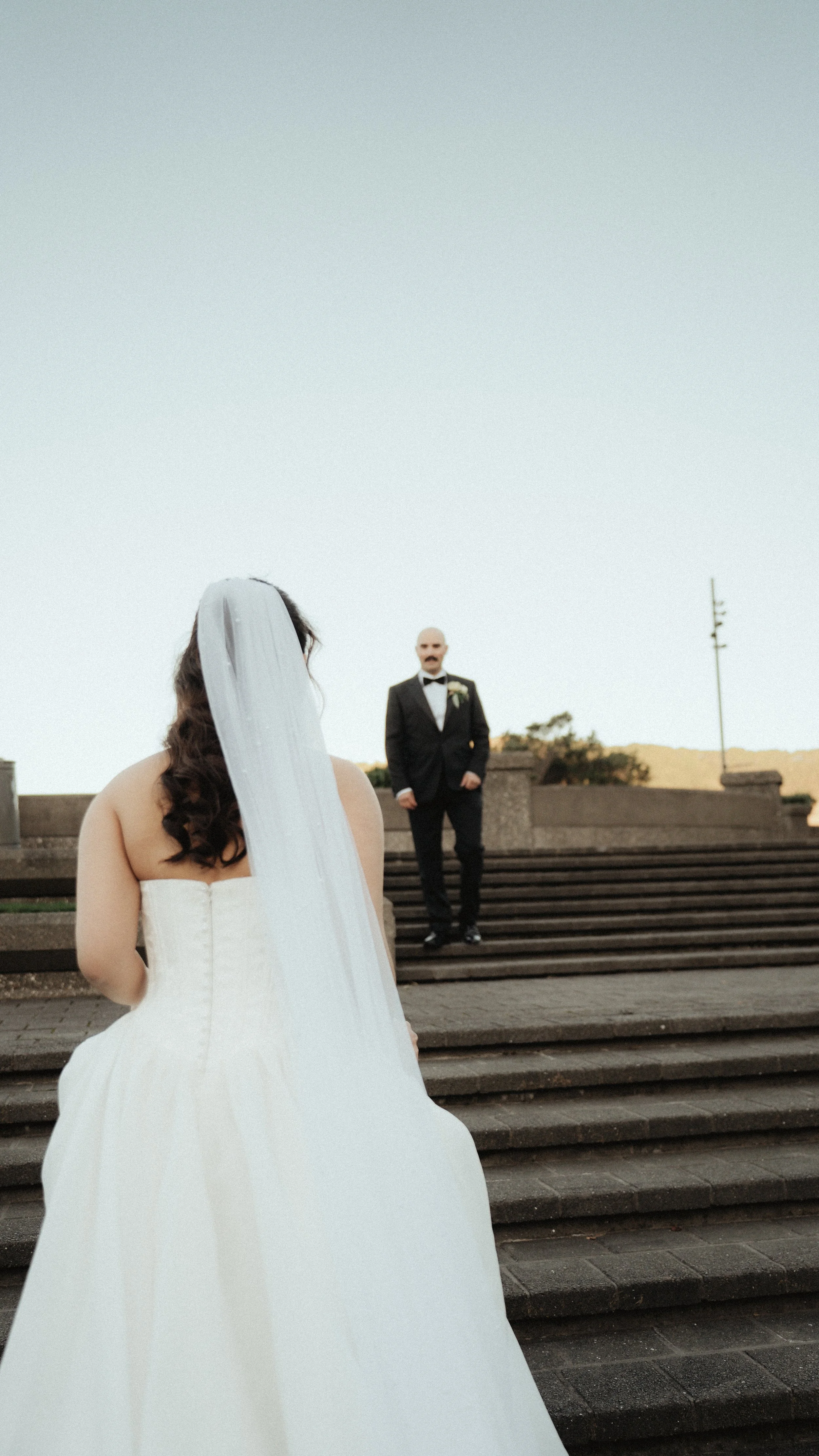 A bride in a white wedding dress and veil stands on steps, facing a groom in a black suit and bow tie, outdoors on a clear day.