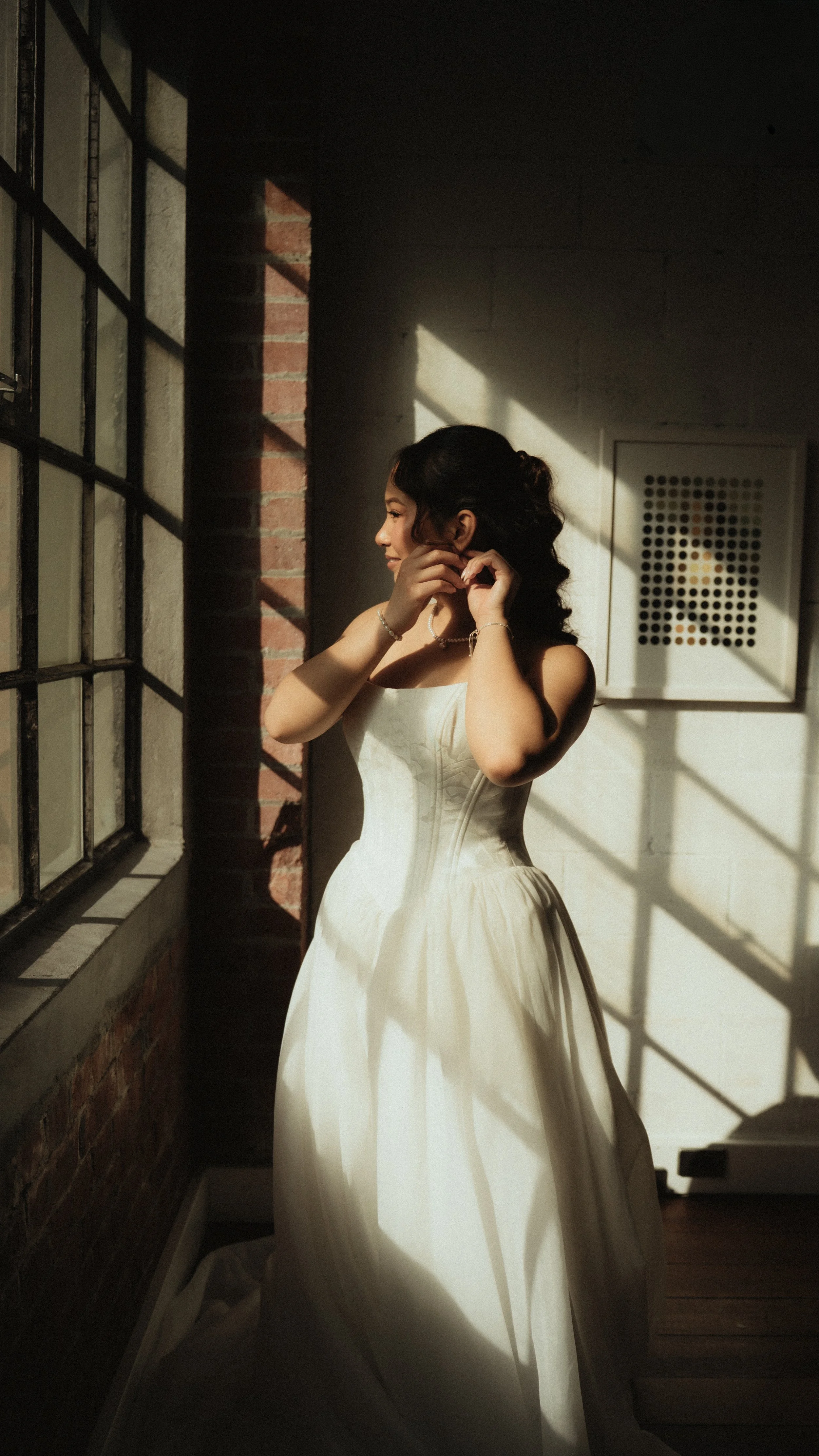 A woman in a white dress standing near a window with sunlight casting shadows, adjusting her earring.
