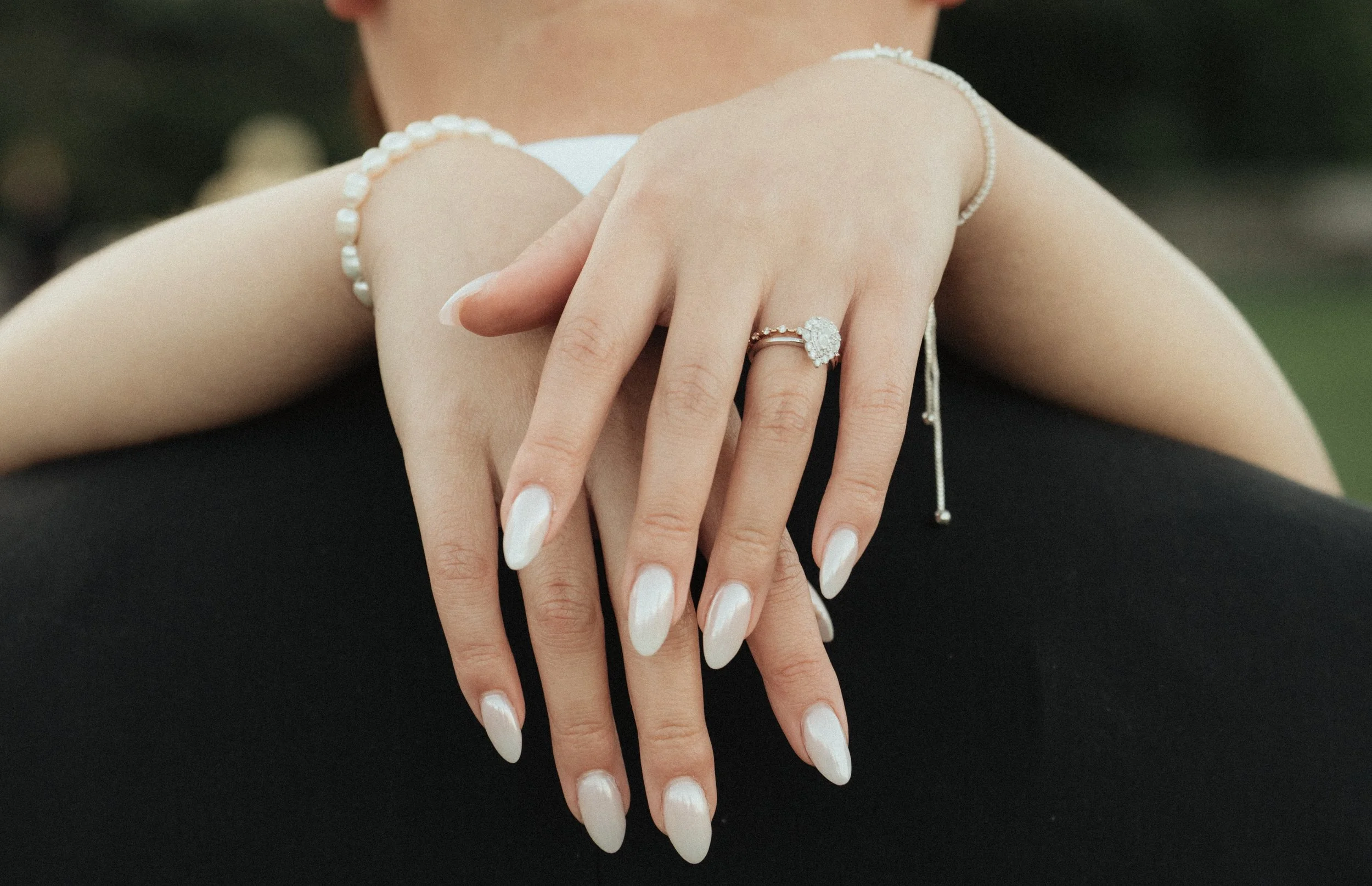 Close-up of a woman's hand displaying a diamond engagement ring and a pearl bracelet, with manicured white nails and blurred background.