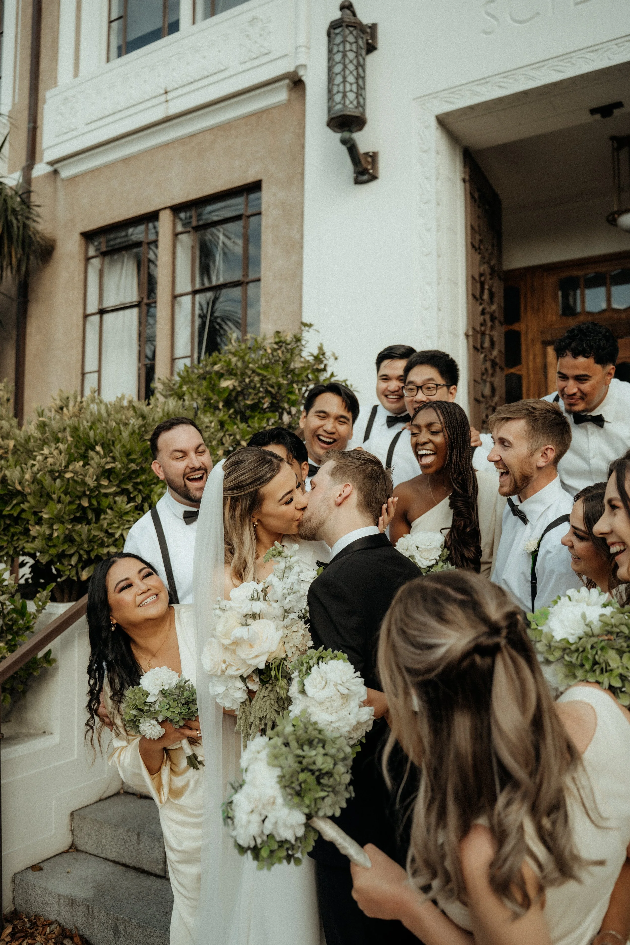 A wedding scene with a bride and groom kissing, surrounded by friends dressed in formal attire. The setting appears to be outside a building with ornate architecture, floral decorations, and people smiling and celebrating.
