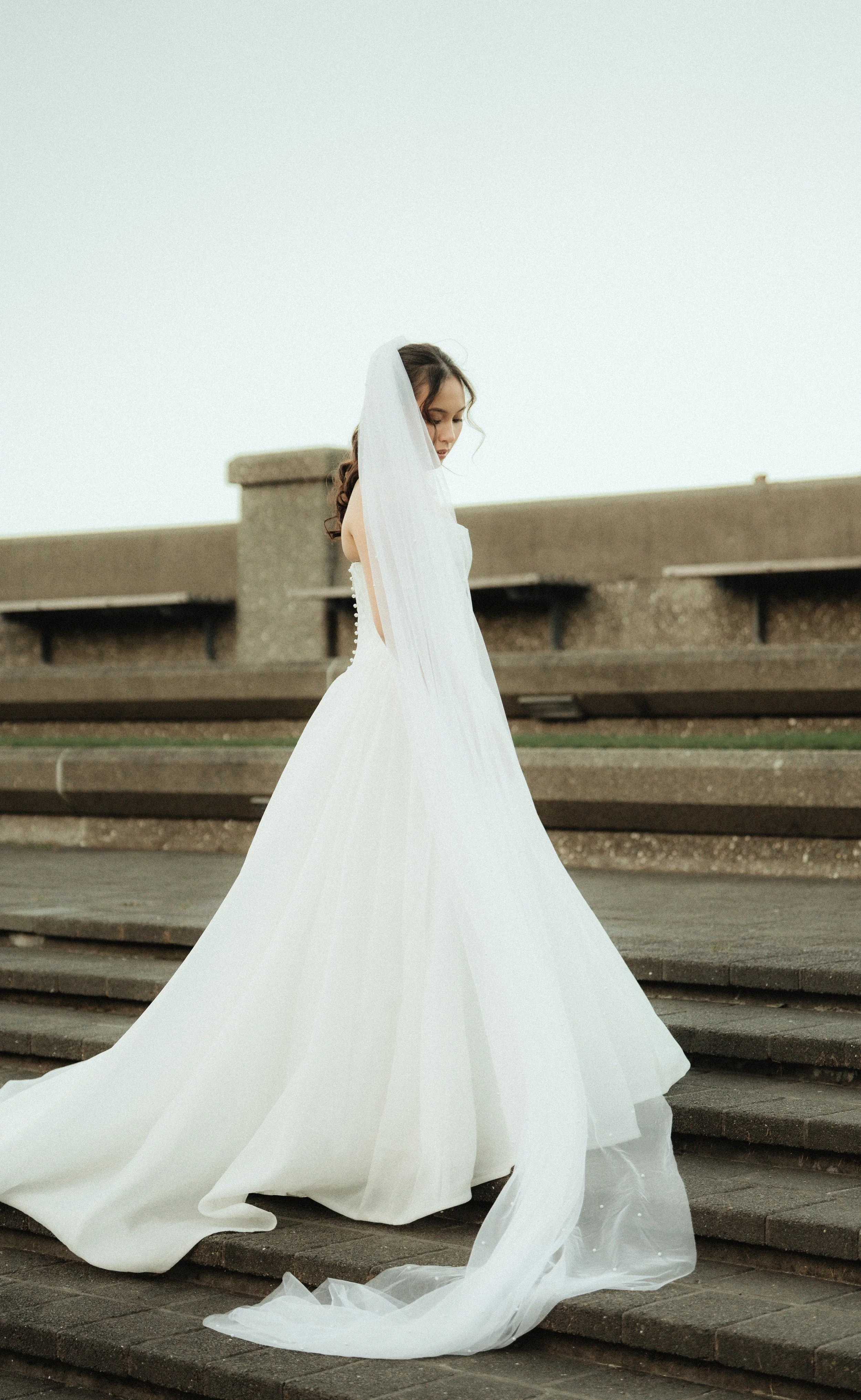 A woman in a white wedding dress with a long train, standing on outdoor concrete steps with a stone railing behind her, looking down and to the side.