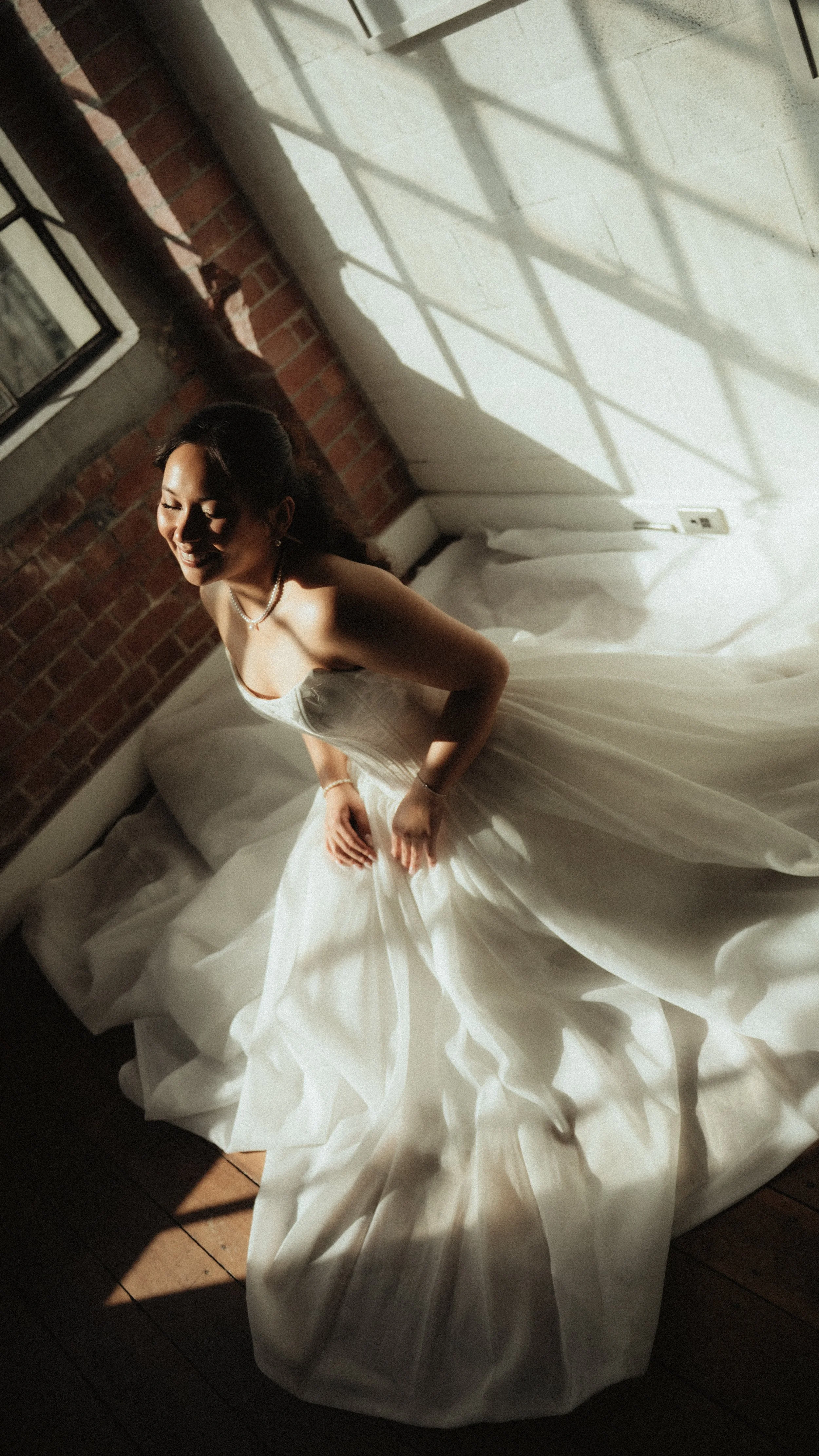 A woman in a white wedding dress is sitting on a bed, smiling with her eyes closed. She is wearing a pearl necklace and earrings, with sunlight casting shadows through a window on her.