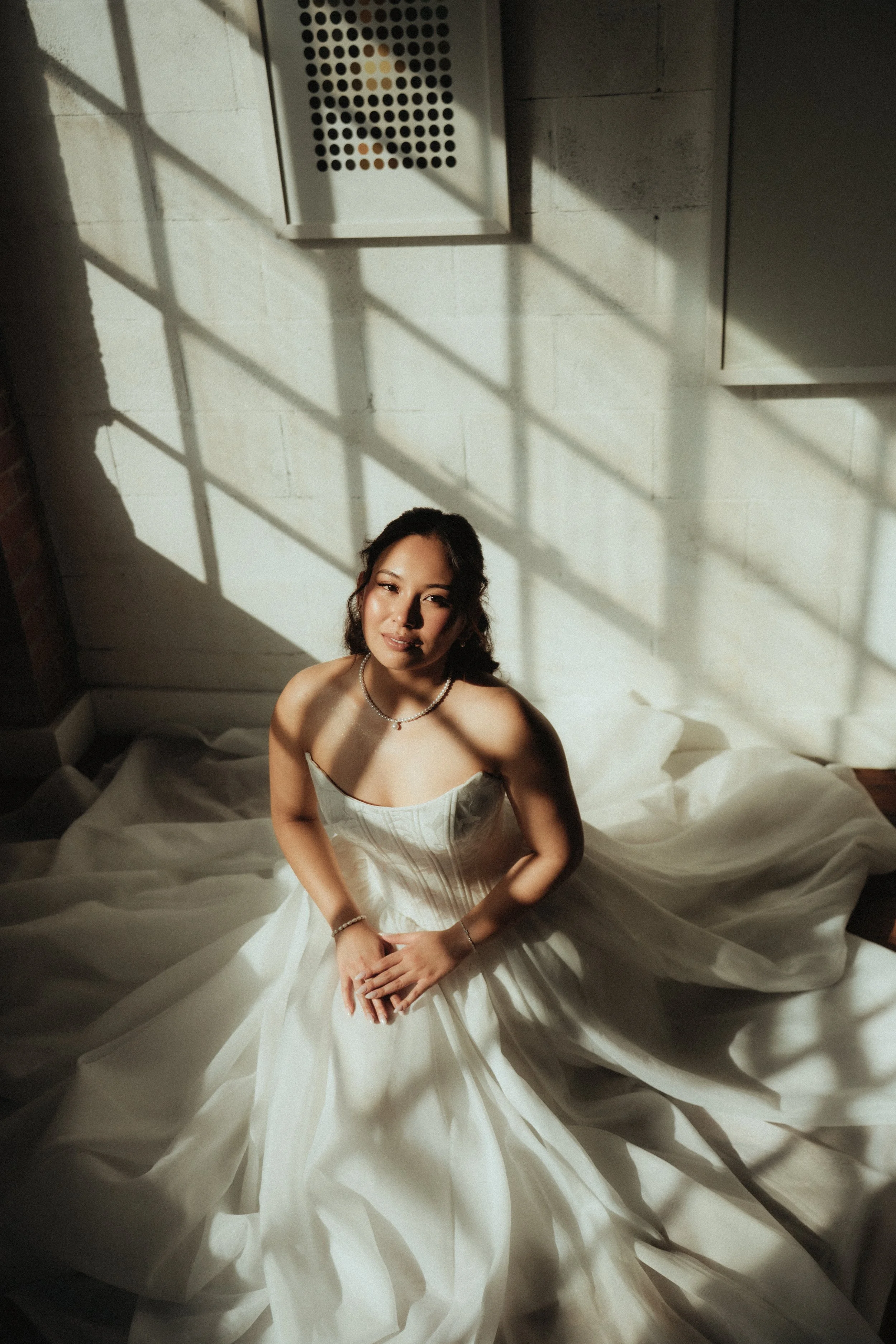 A woman in a white dress sitting on a bed with sunlight casting shadows through a window, creating a grid pattern on the wall and floor.