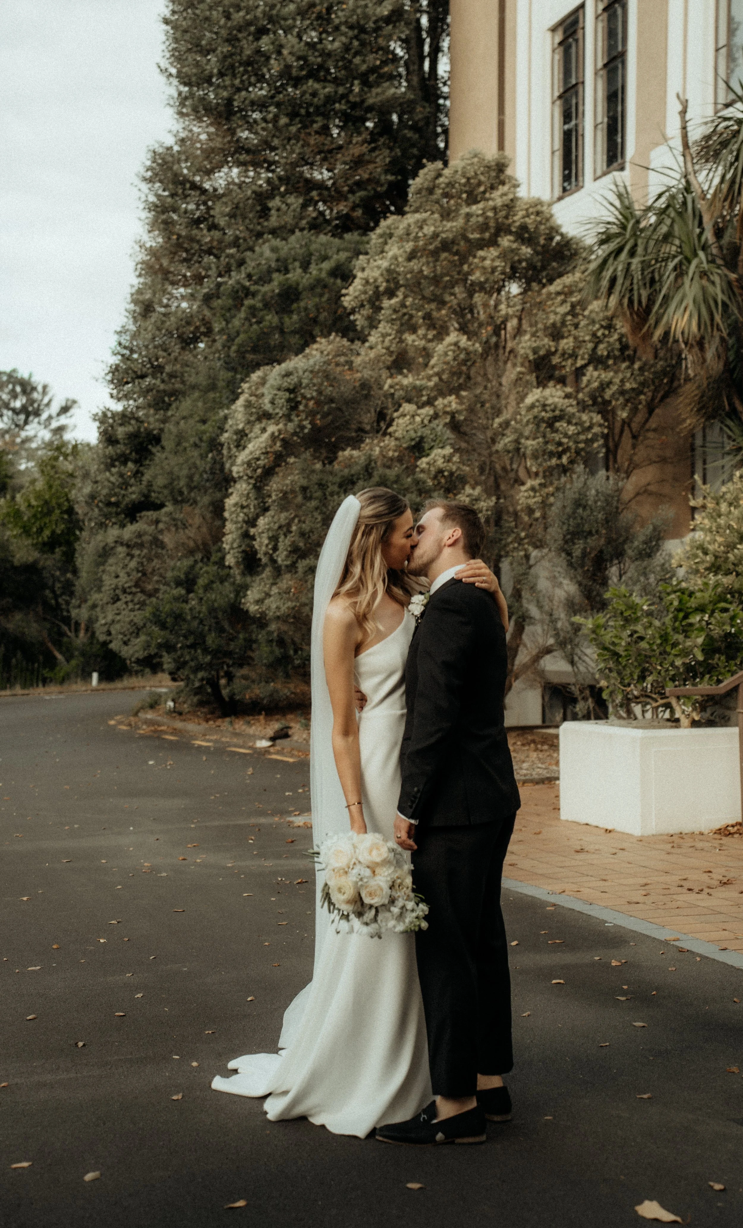 A bride and groom sharing a kiss outdoors during their wedding, with the bride holding a bouquet of white roses and the groom in a black tuxedo. There are trees and a building in the background.