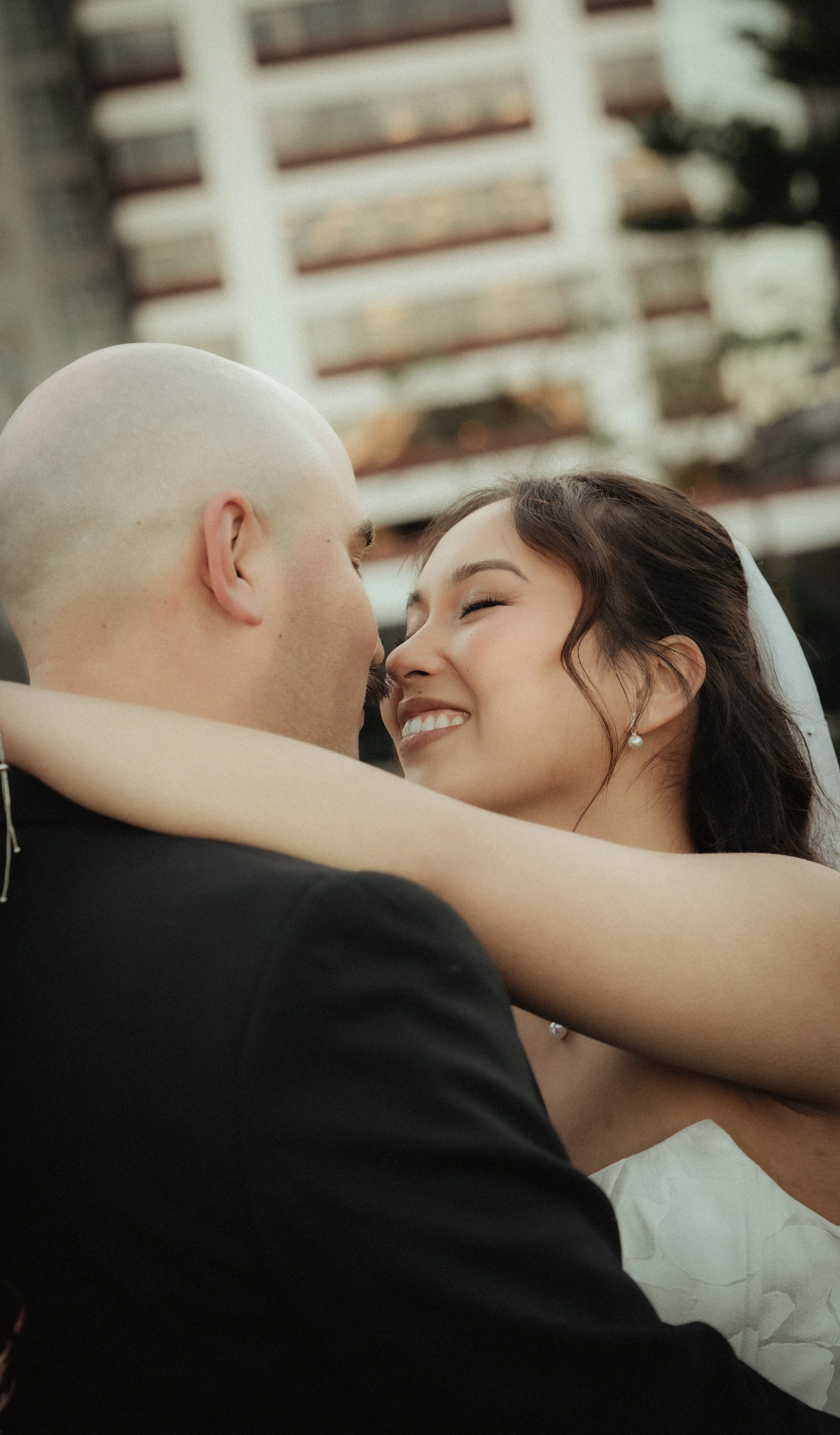 A couple embracing outdoors, with the man in a black suit and the woman in a white dress, smiling and looking at each other, with a building in the background.