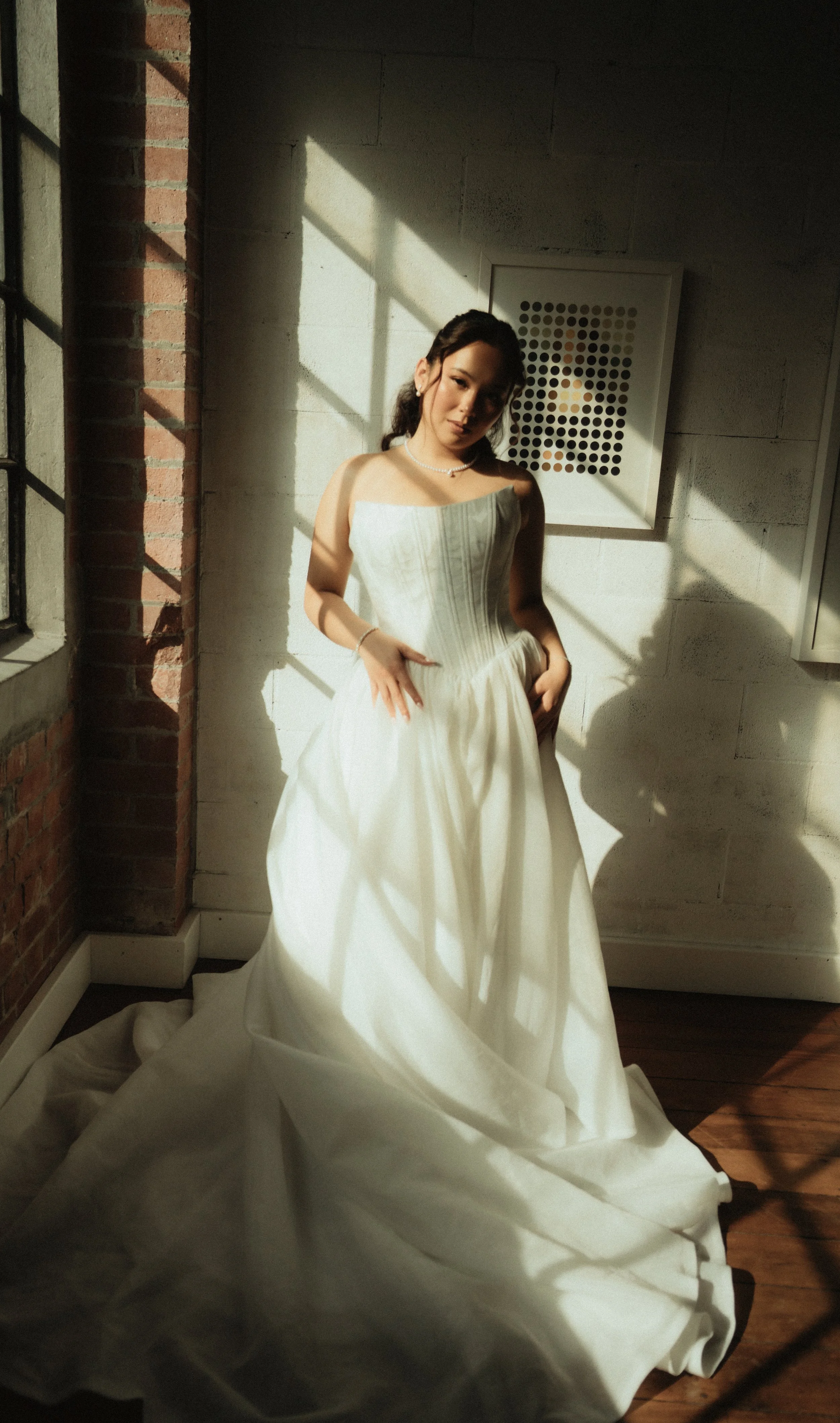 A woman in a white strapless wedding gown standing in a room with brick and concrete walls, sunlight casting shadows on her and the wall behind her, with a framed piece of art on the wall.