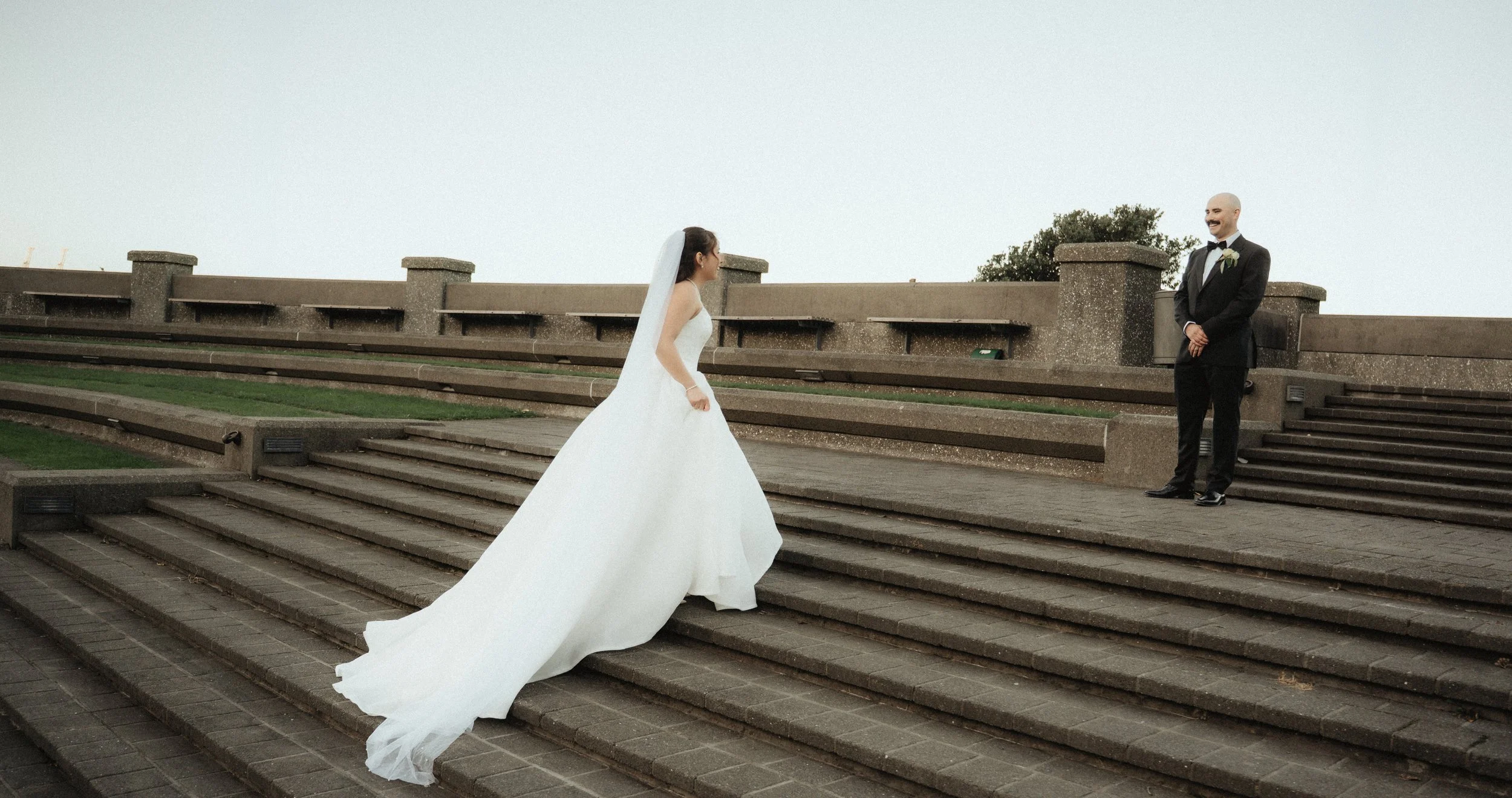 A bride in a white wedding gown standing on stairs, facing a groom in a tuxedo, who is smiling at her against a gray sky background.
