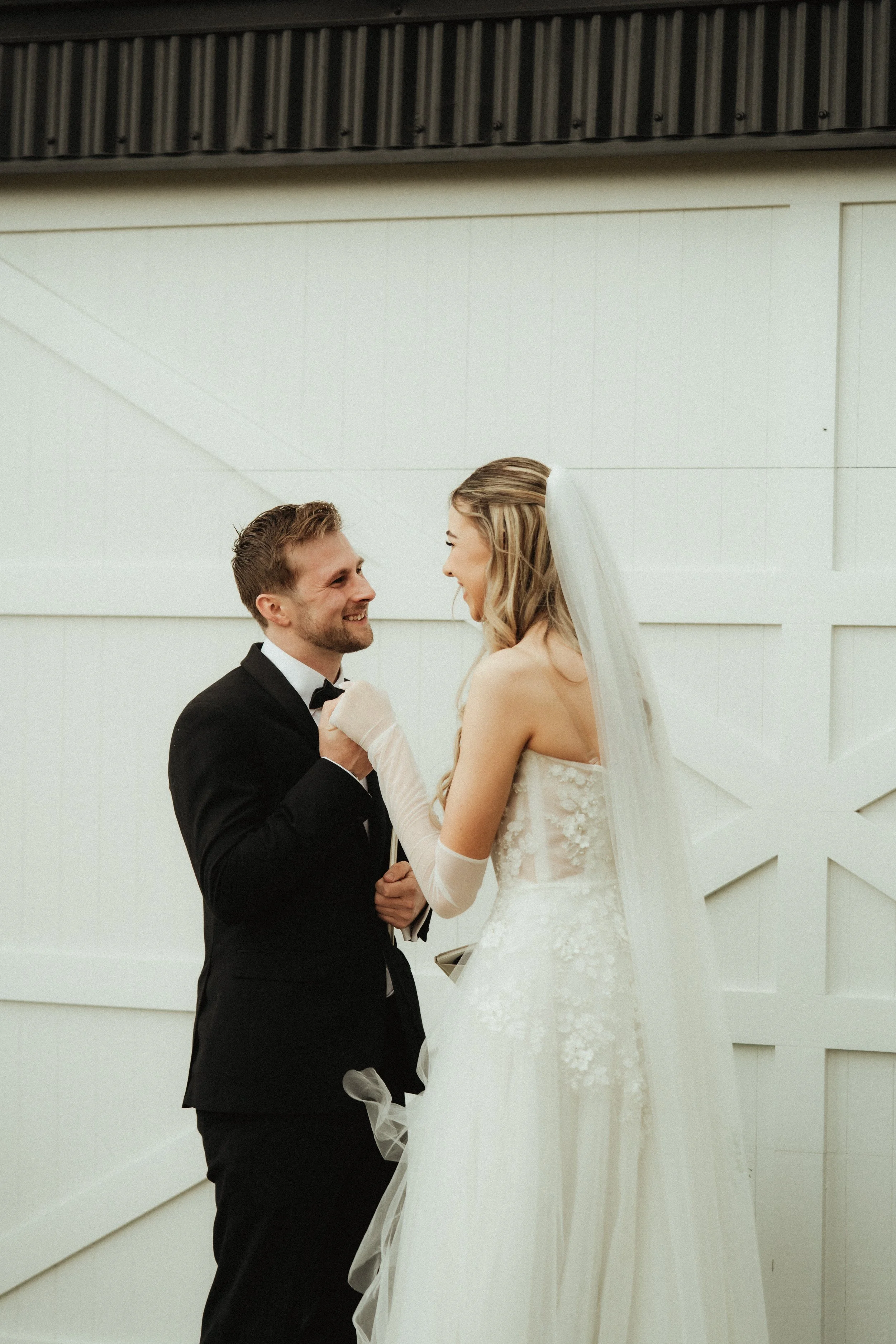 A bride and groom share a moment of joy during their wedding ceremony. The groom is wearing a black tuxedo with a bow tie, and the bride is dressed in a white wedding gown with a veil. They are holding hands and smiling at each other against a simple
